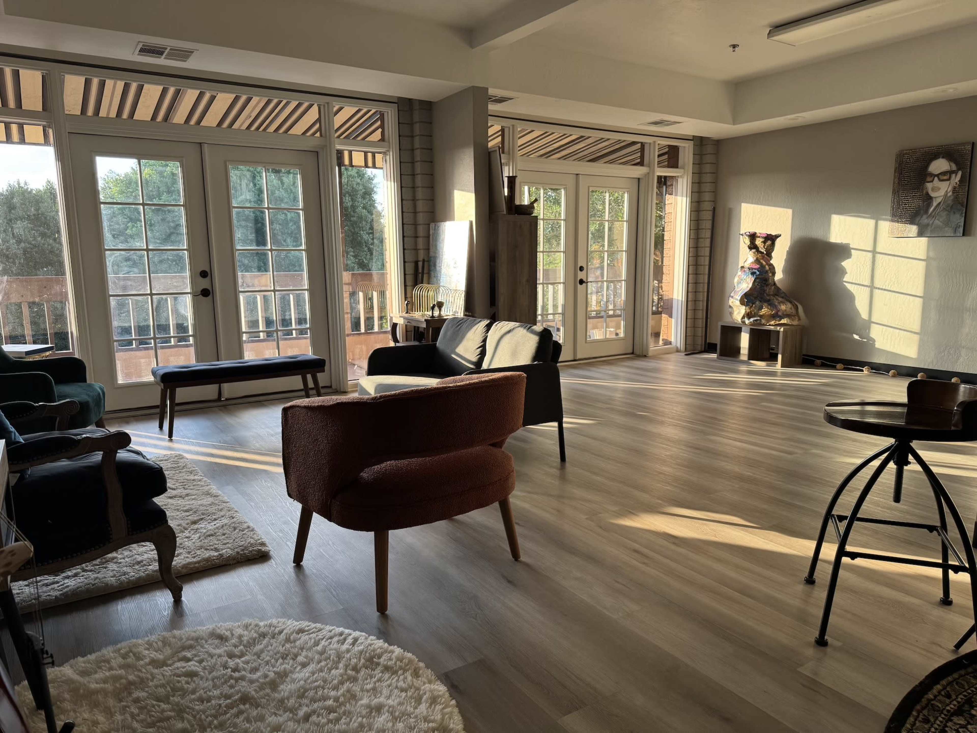 The image shows a spacious living area with large windows, furnished with various seating options, and wooden flooring.