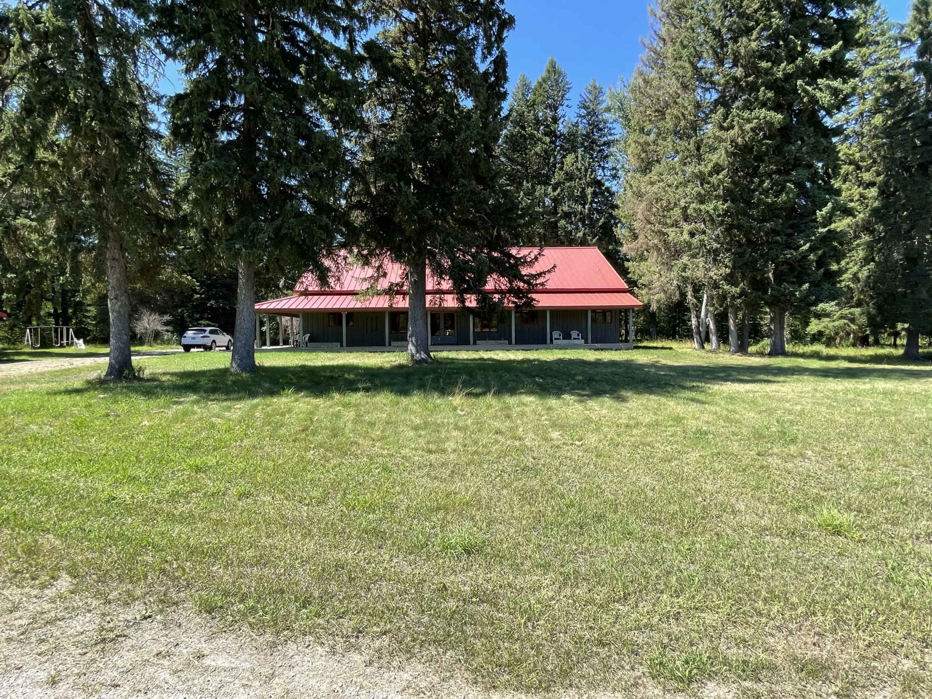 A single-story house with a red metal roof is surrounded by tall trees and green grass in a rural setting.