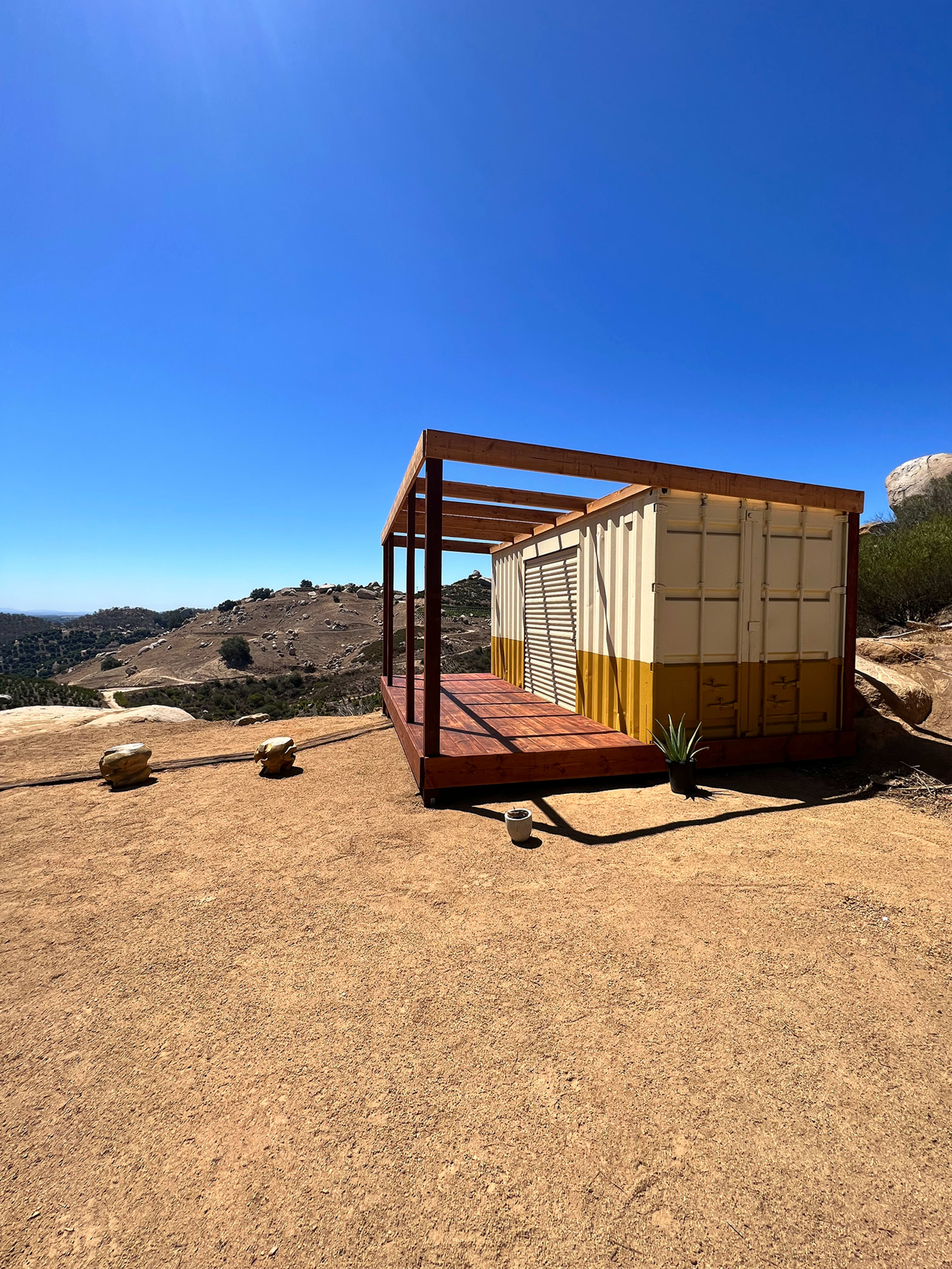 A modern structure made from shipping containers is positioned on a sandy hillside with a clear blue sky in the background.
