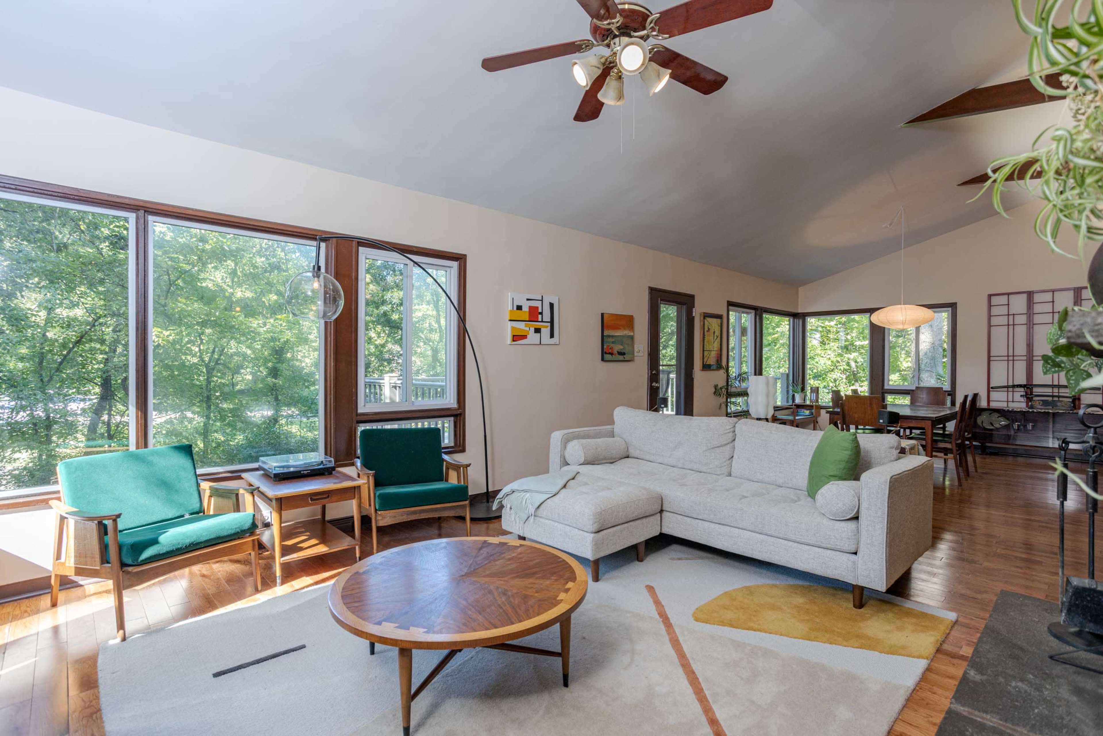 A spacious living room with large windows, a light-colored sofa, a round coffee table, and green chairs, surrounded by greenery outside.