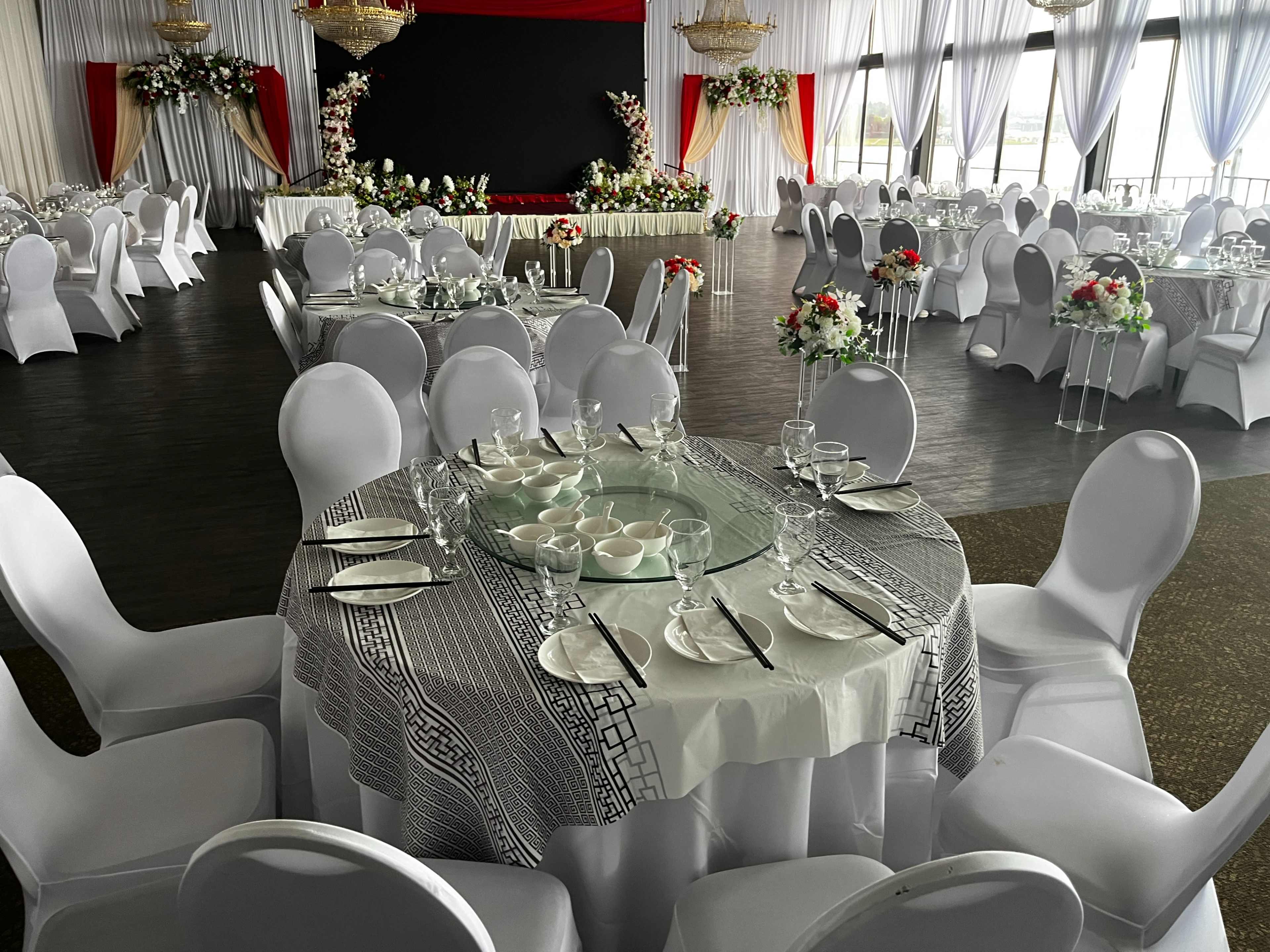 A spacious banquet hall is set up for an event, featuring round tables with white tablecloths, elegant place settings, and floral centerpieces, along with a stage at the front.