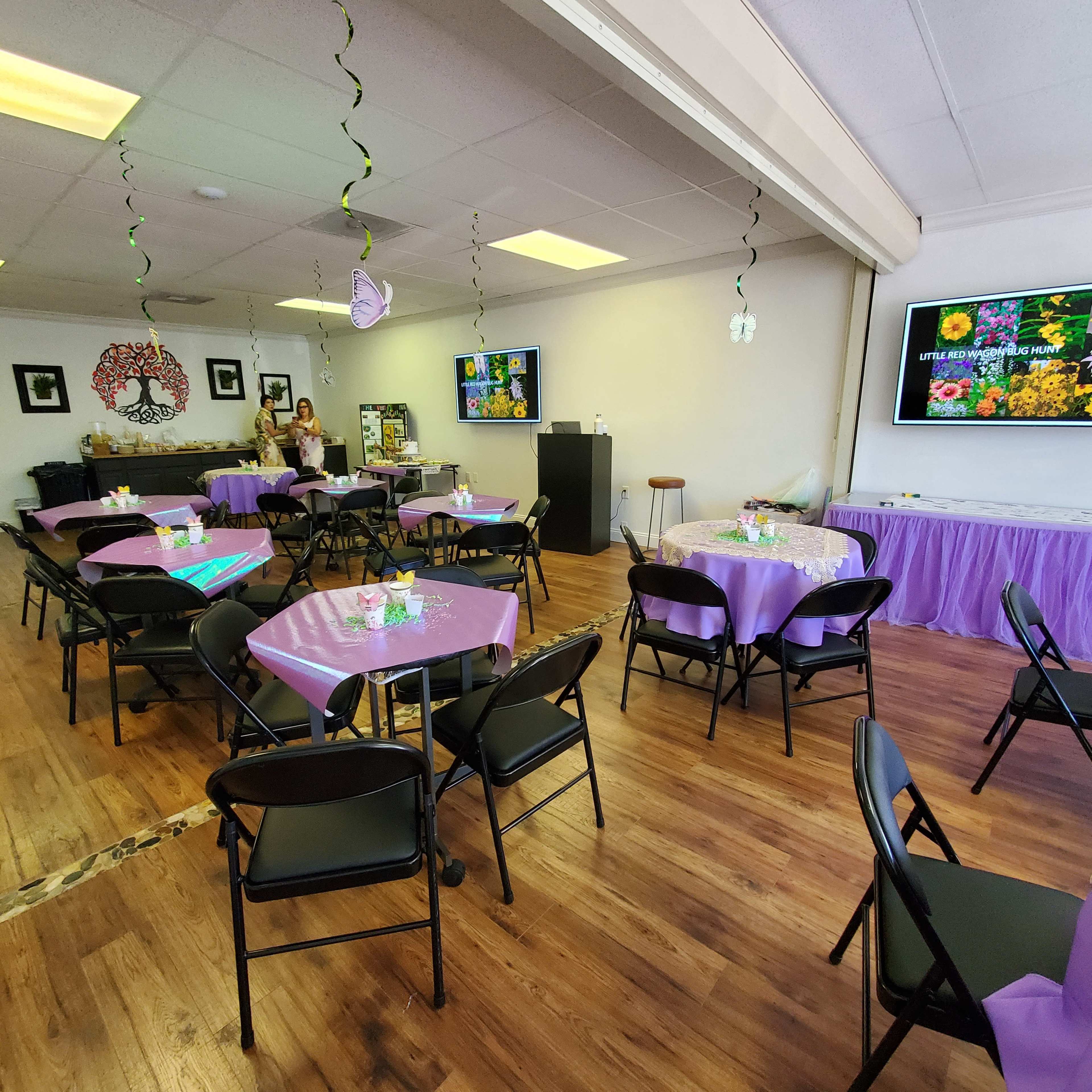 The image shows a brightly decorated event space with purple tablecloths, chairs arranged around tables, and screens displaying floral images.