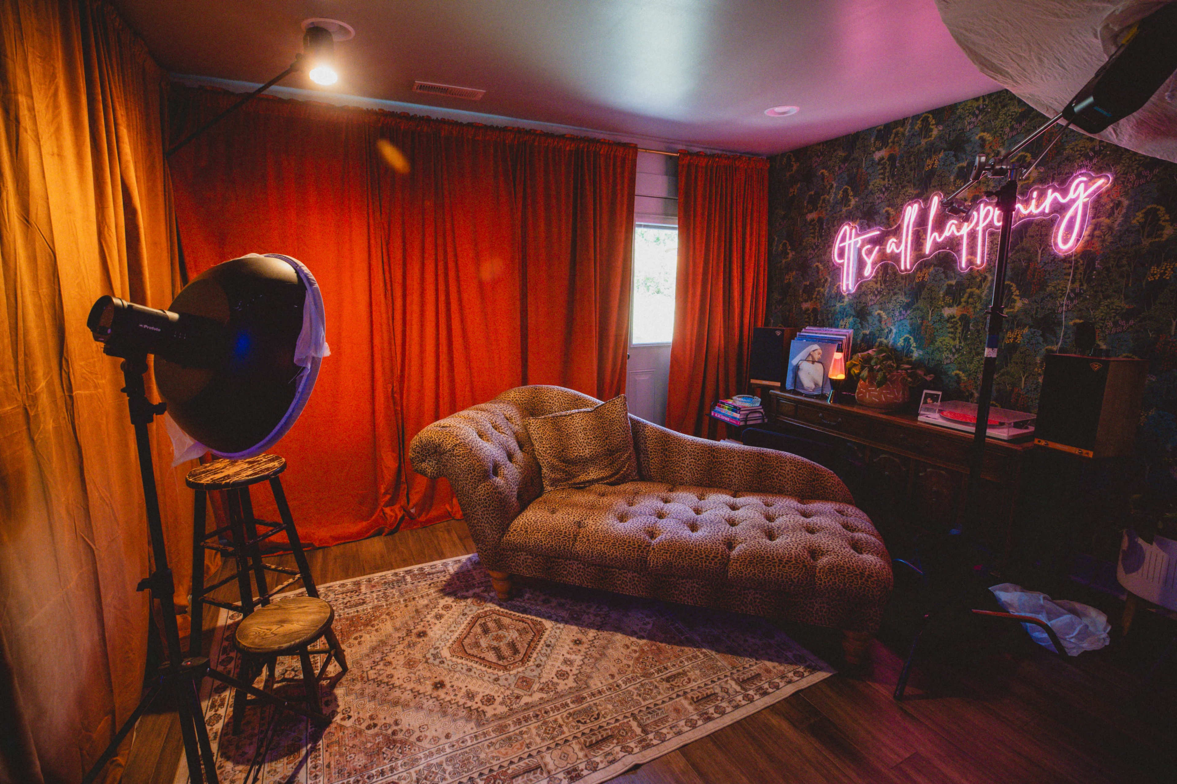 The image depicts a cozy room featuring a tufted chaise lounge, a brightly lit neon sign that reads "It’s all happening," and a vintage wooden side table against a decorative wall covered in patterned wallpaper.