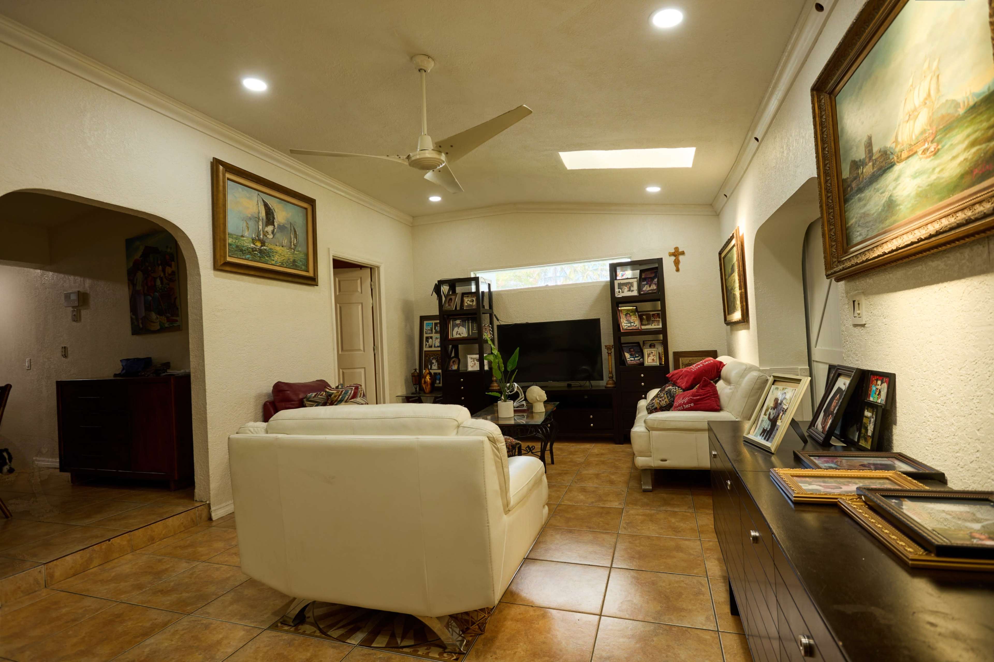 The image shows a living room with a ceiling fan, a television on a black stand, two white sofas, and framed pictures on the walls.