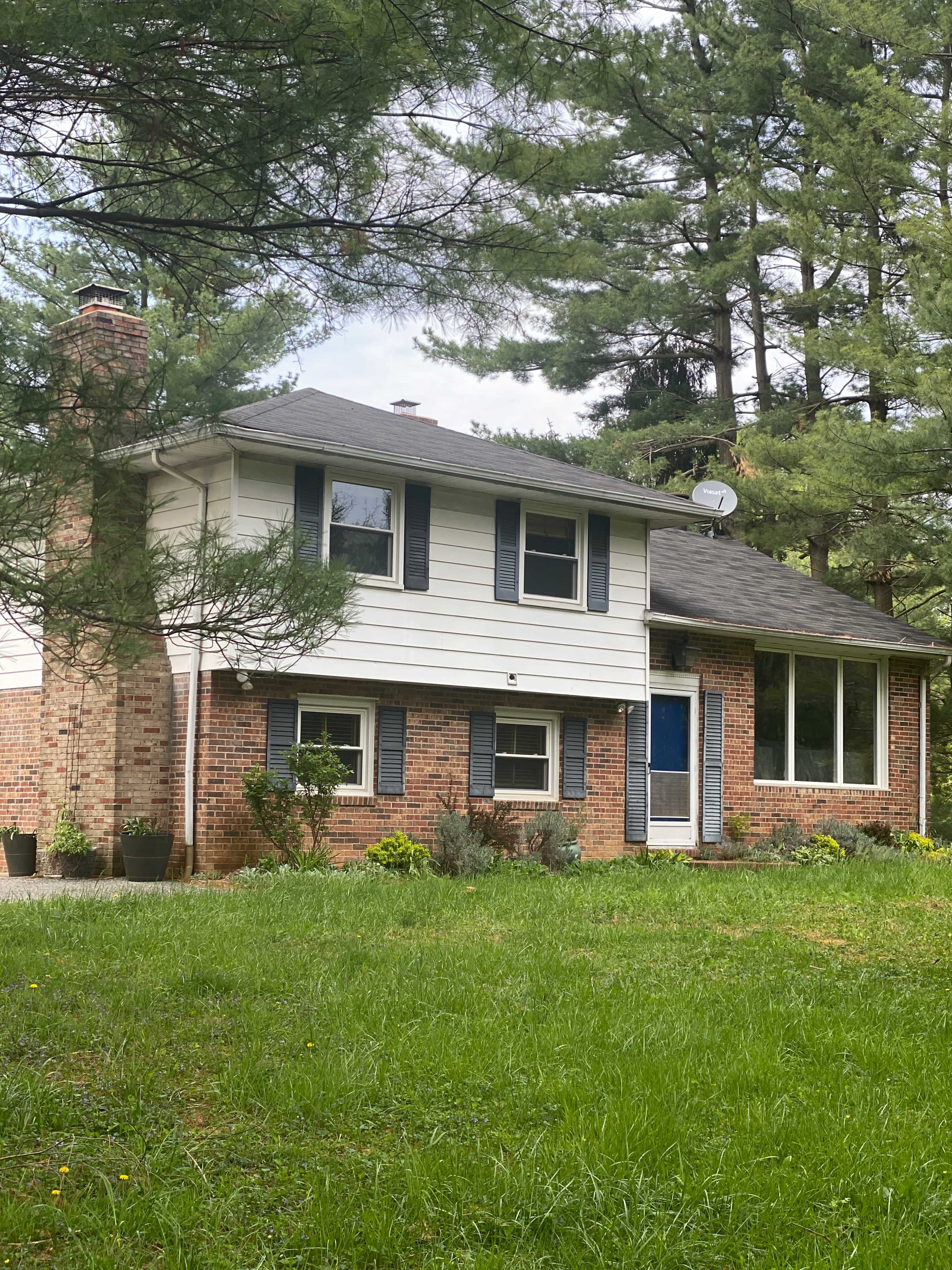 A two-story house with a brick and siding exterior is surrounded by tall pine trees and a grassy lawn.