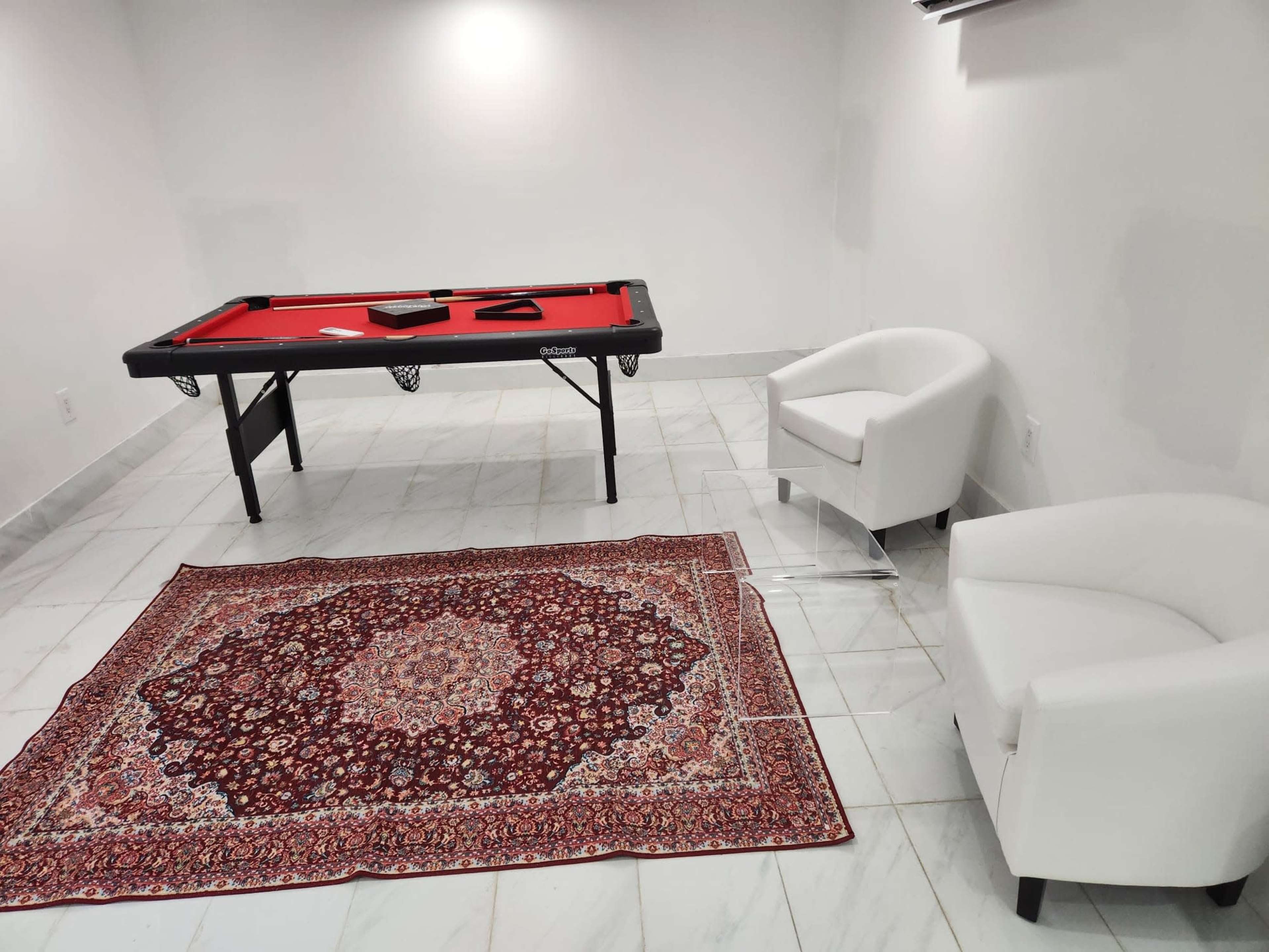 A pool table with a red felt surface is set up in a room featuring white walls and two white armchairs placed around a decorative rug on the floor.
