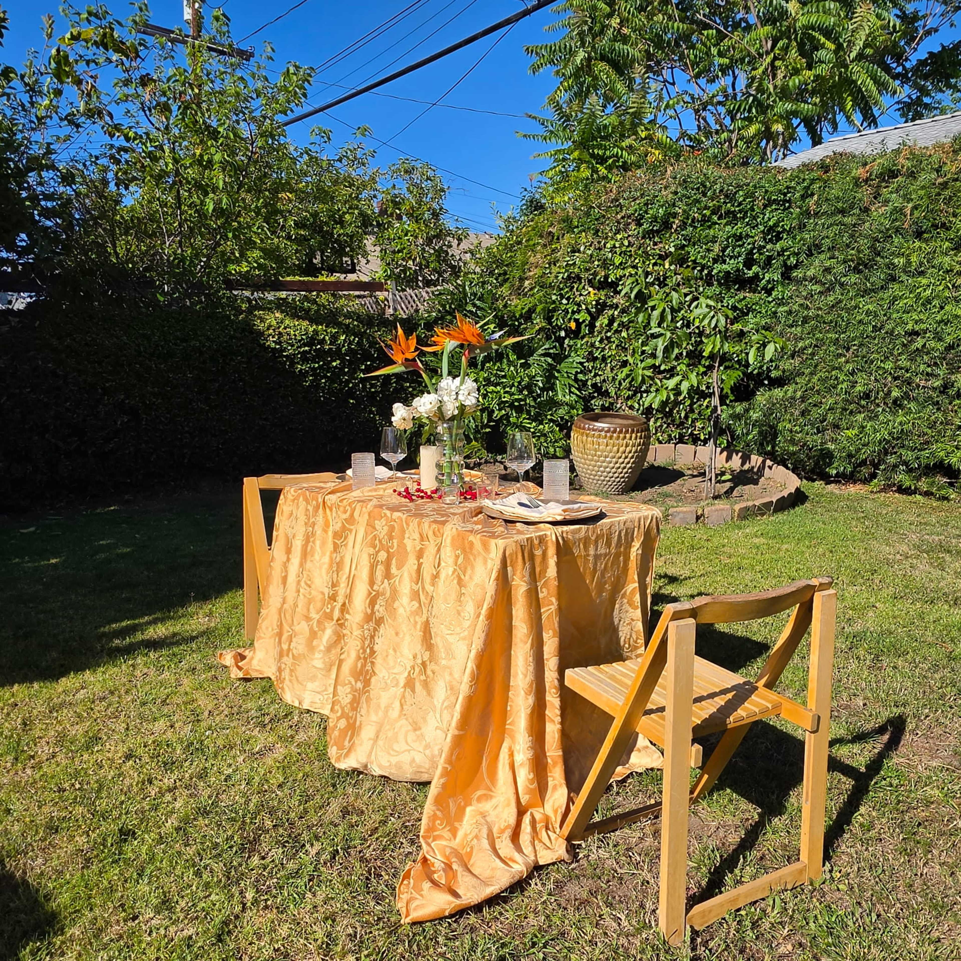 An elegantly set outdoor dining table with a golden tablecloth, surrounded by greenery and a clear blue sky.