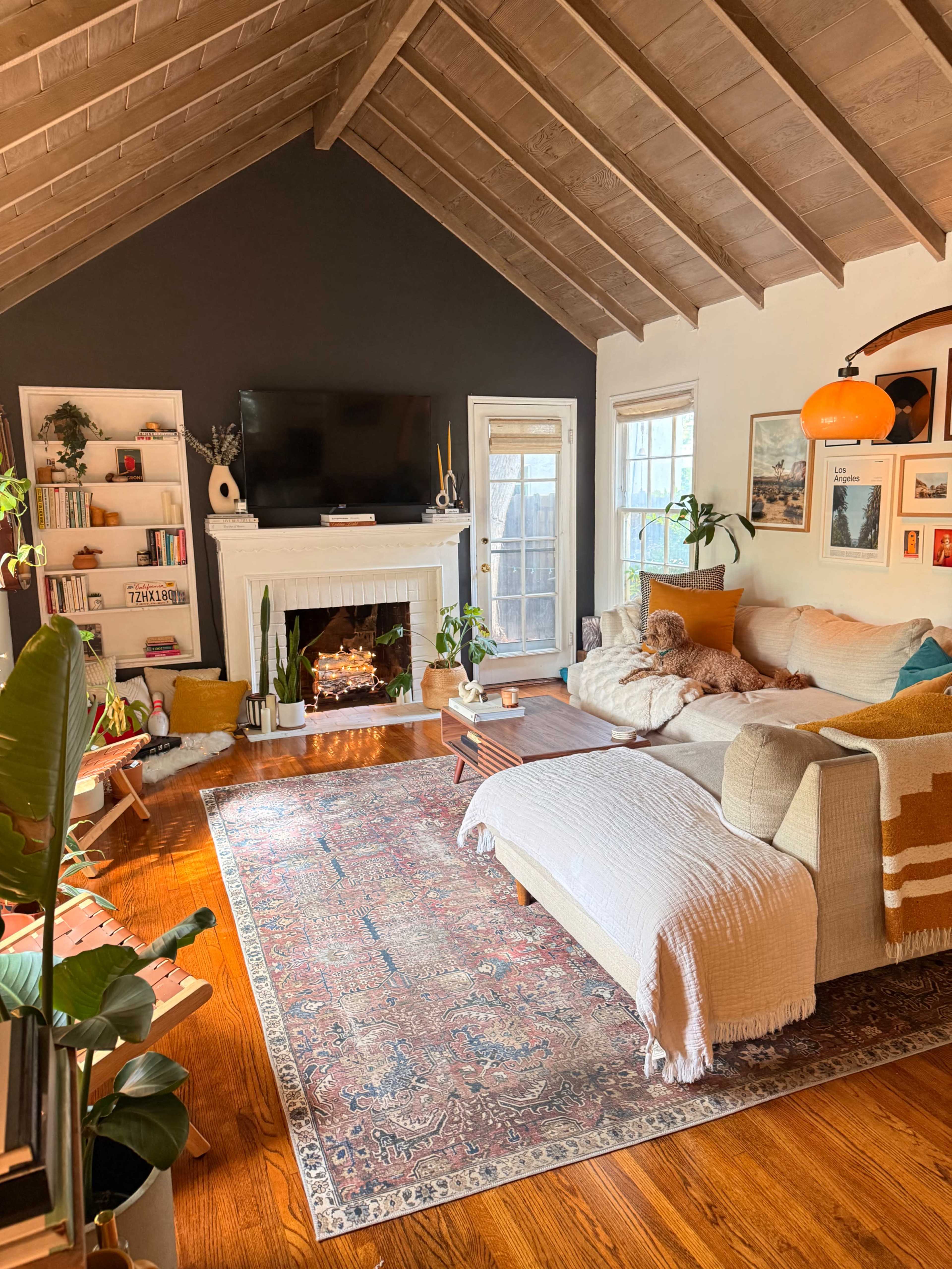The cozy living room features a high, wooden ceiling, a fireplace with a TV above, and comfortable furnishings, accented by a large area rug and shelves filled with books and decorative items.