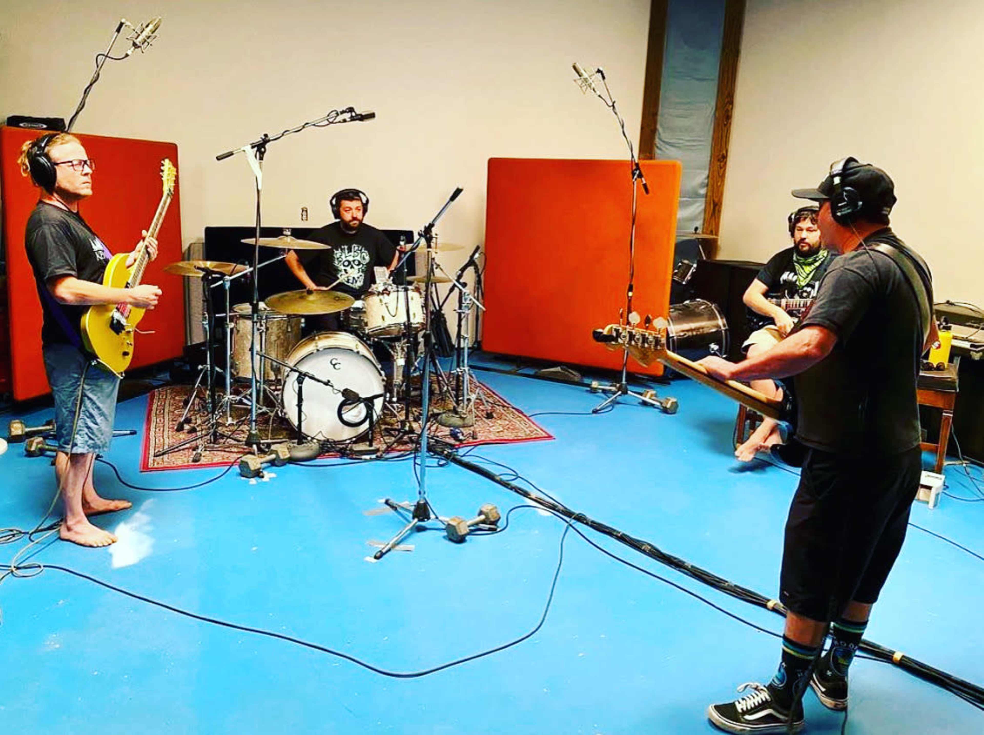 A group of four musicians is playing instruments in a sound studio with orange soundproofing panels and microphones set up around them.