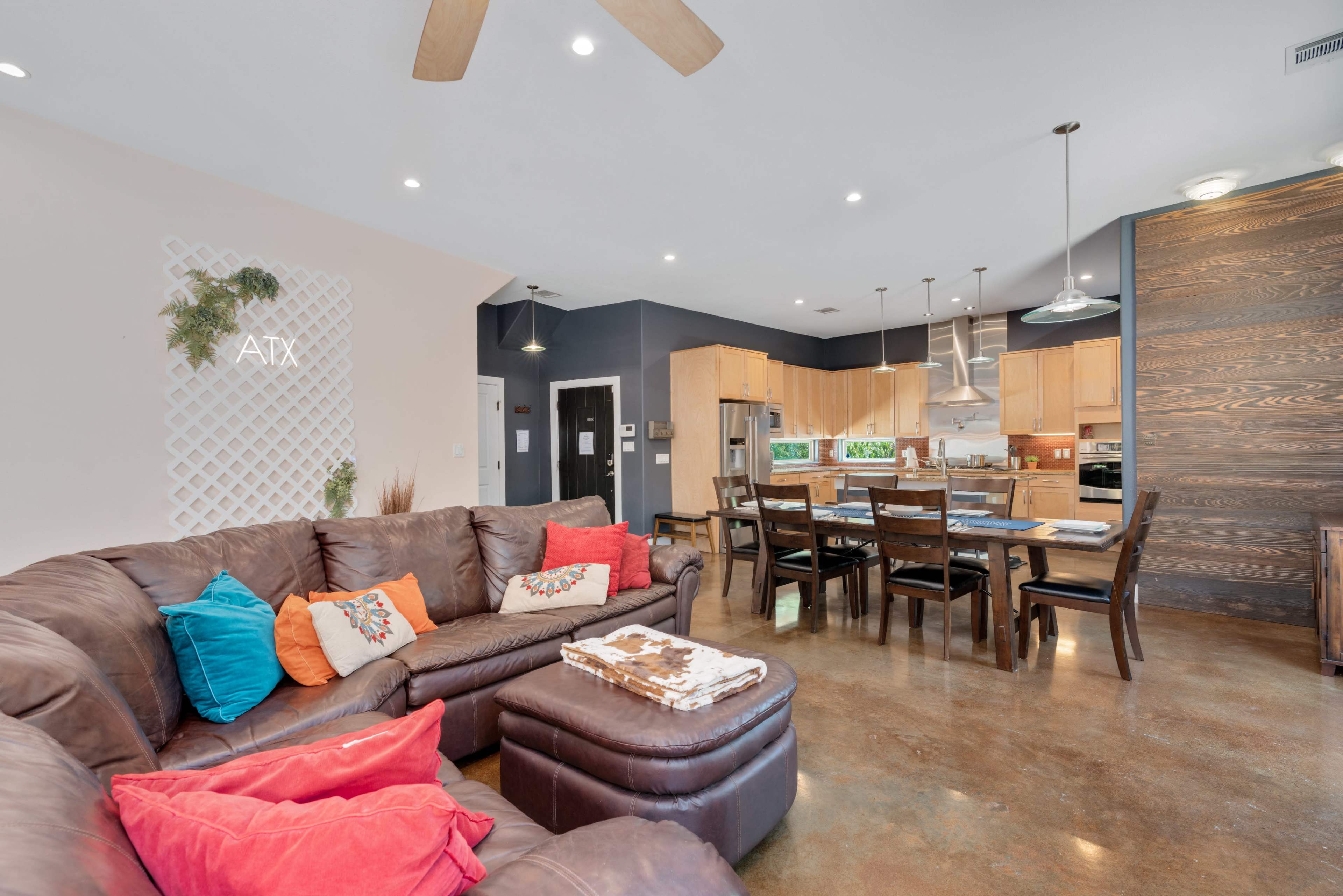A spacious living area features a brown sectional sofa with colorful cushions, a dining table, and a modern kitchen with wooden cabinets and stainless steel appliances.