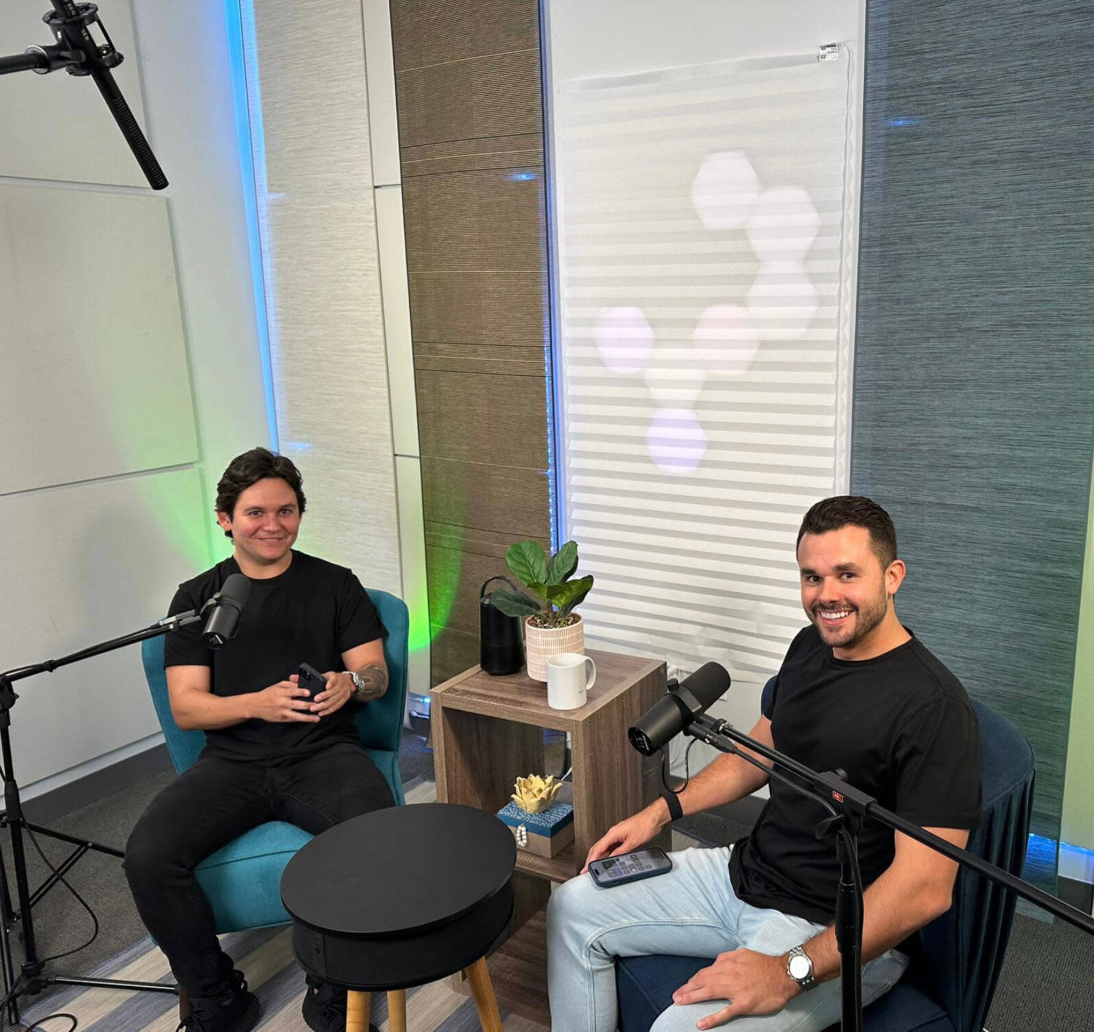 Two men sit in a modern studio setting with microphones in front of them, surrounded by plants and a small table.