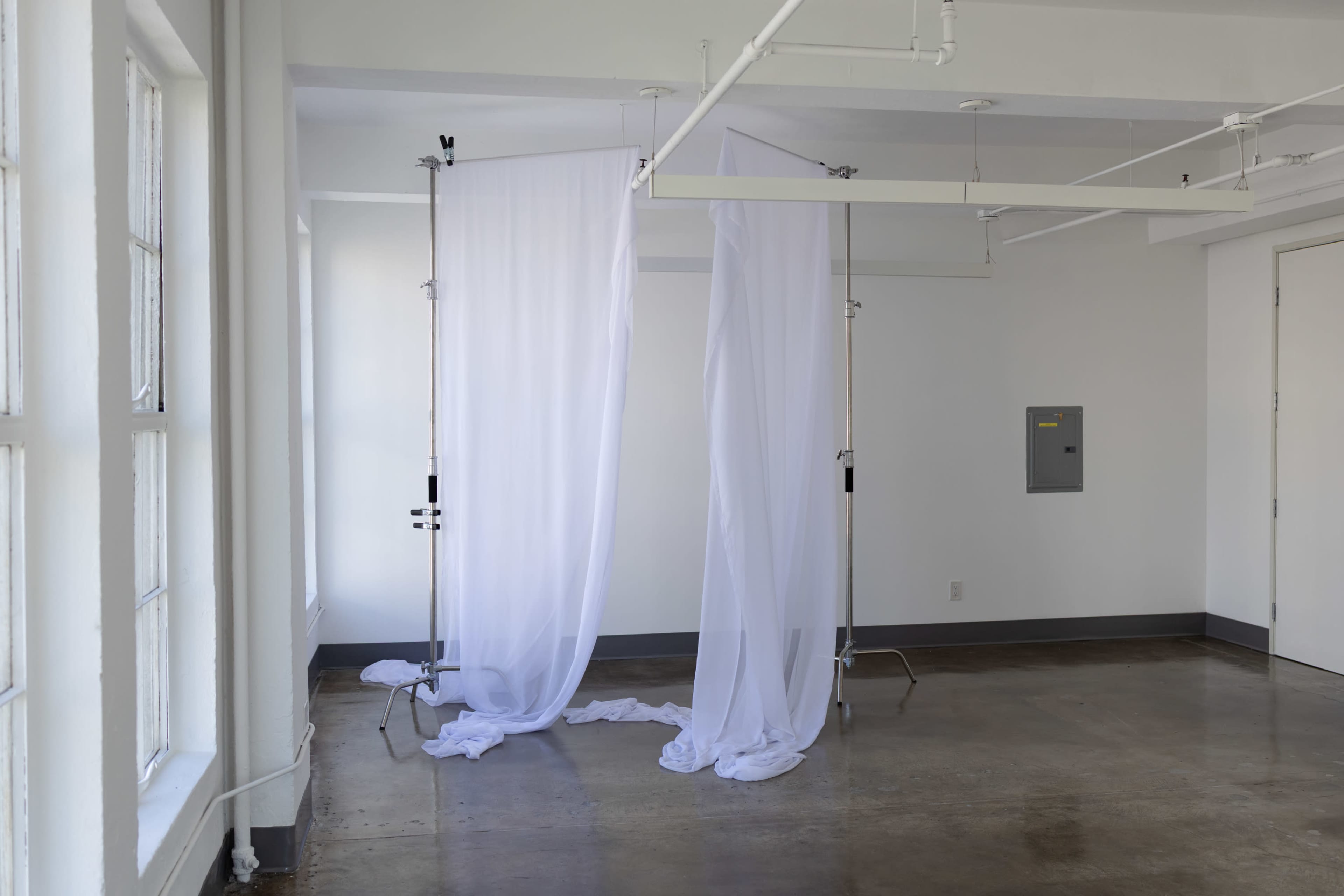 The image features two sheer white curtains draped between tall stands in a minimalist, well-lit room with white walls and a concrete floor.
