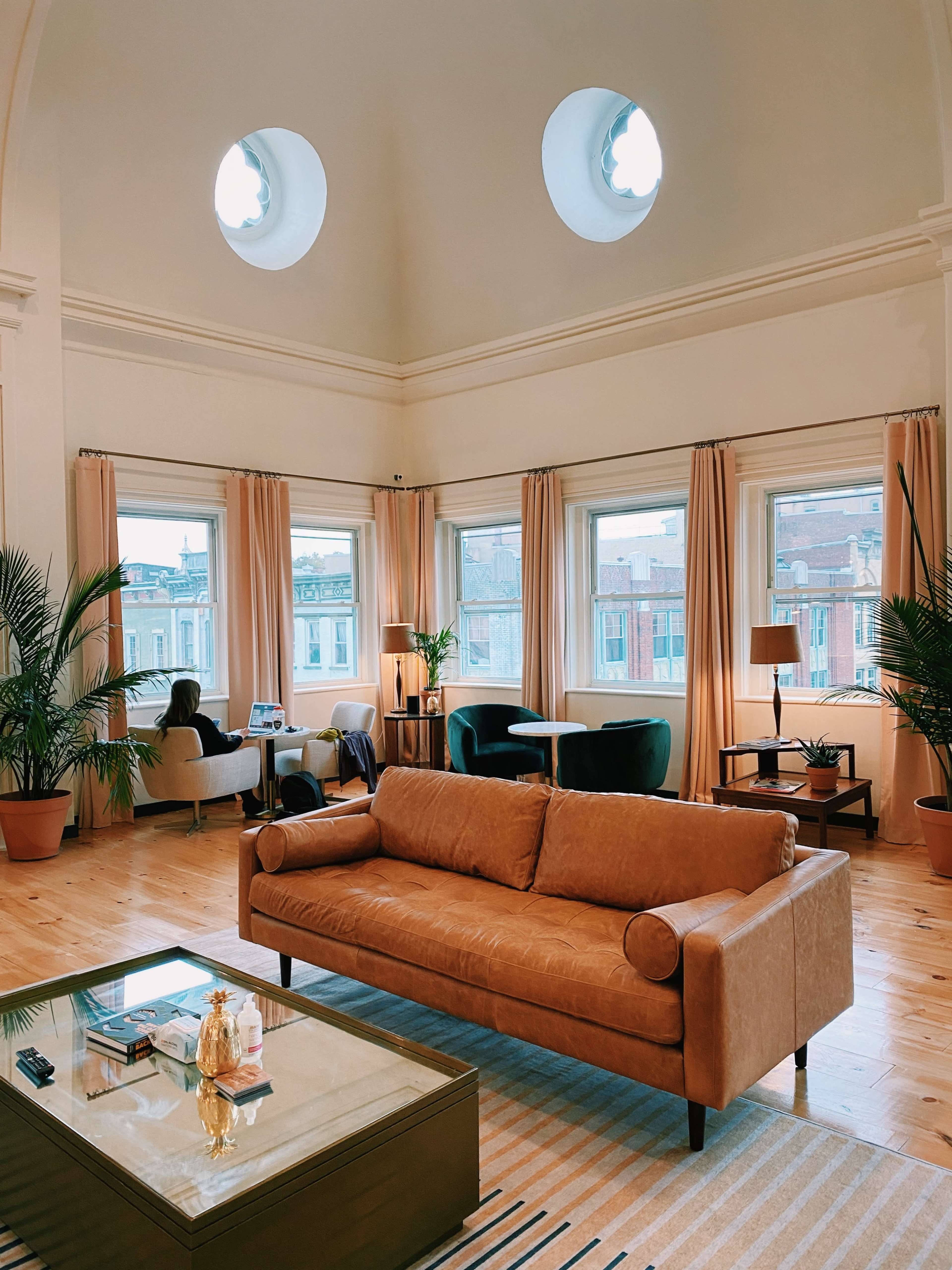 A spacious living room features a brown leather sofa, large windows with light curtains, and modern furnishings.