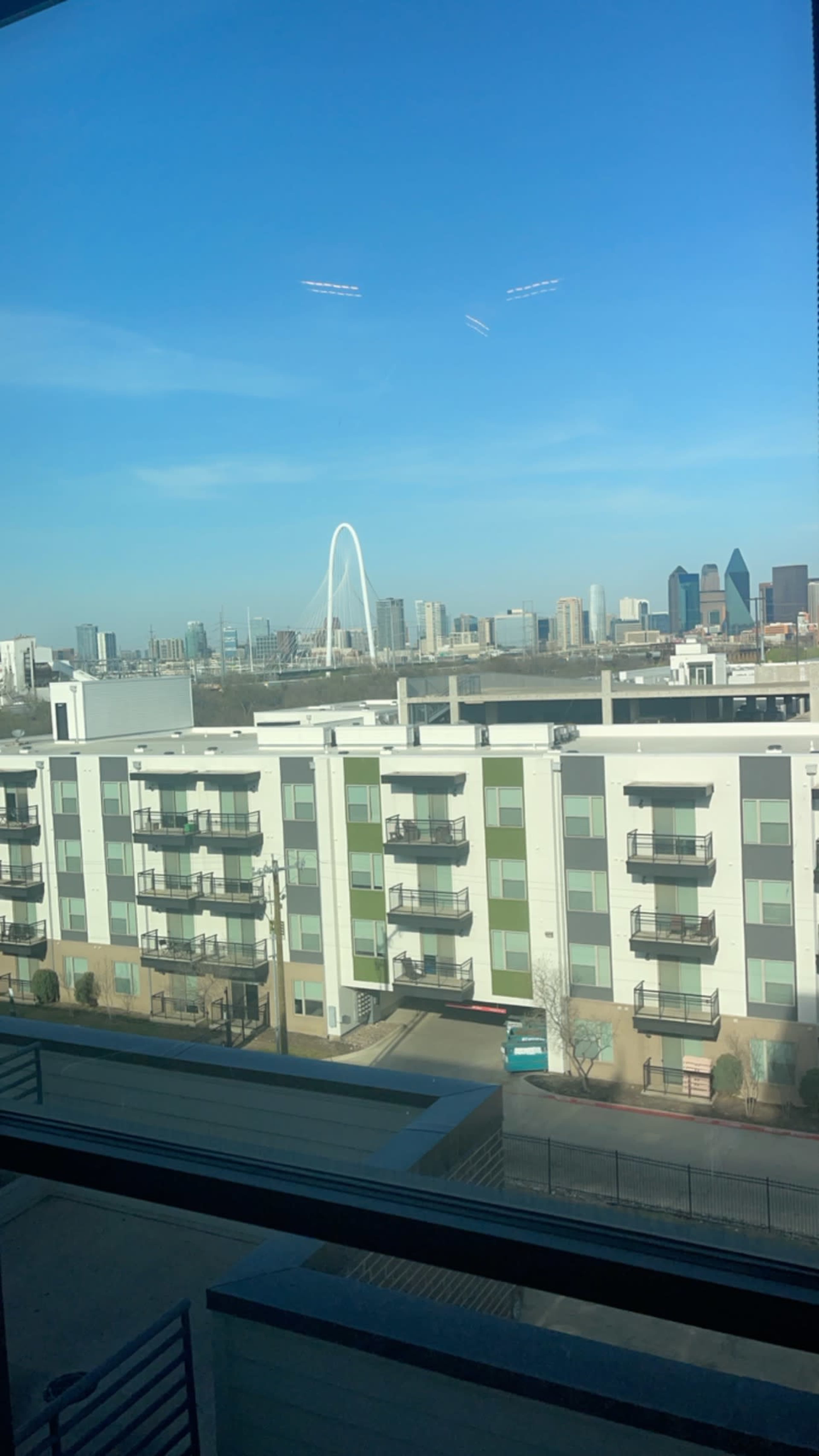 The image shows a view of modern apartment buildings in the foreground with a city skyline, including a distinctive arch bridge, in the background under a clear blue sky.