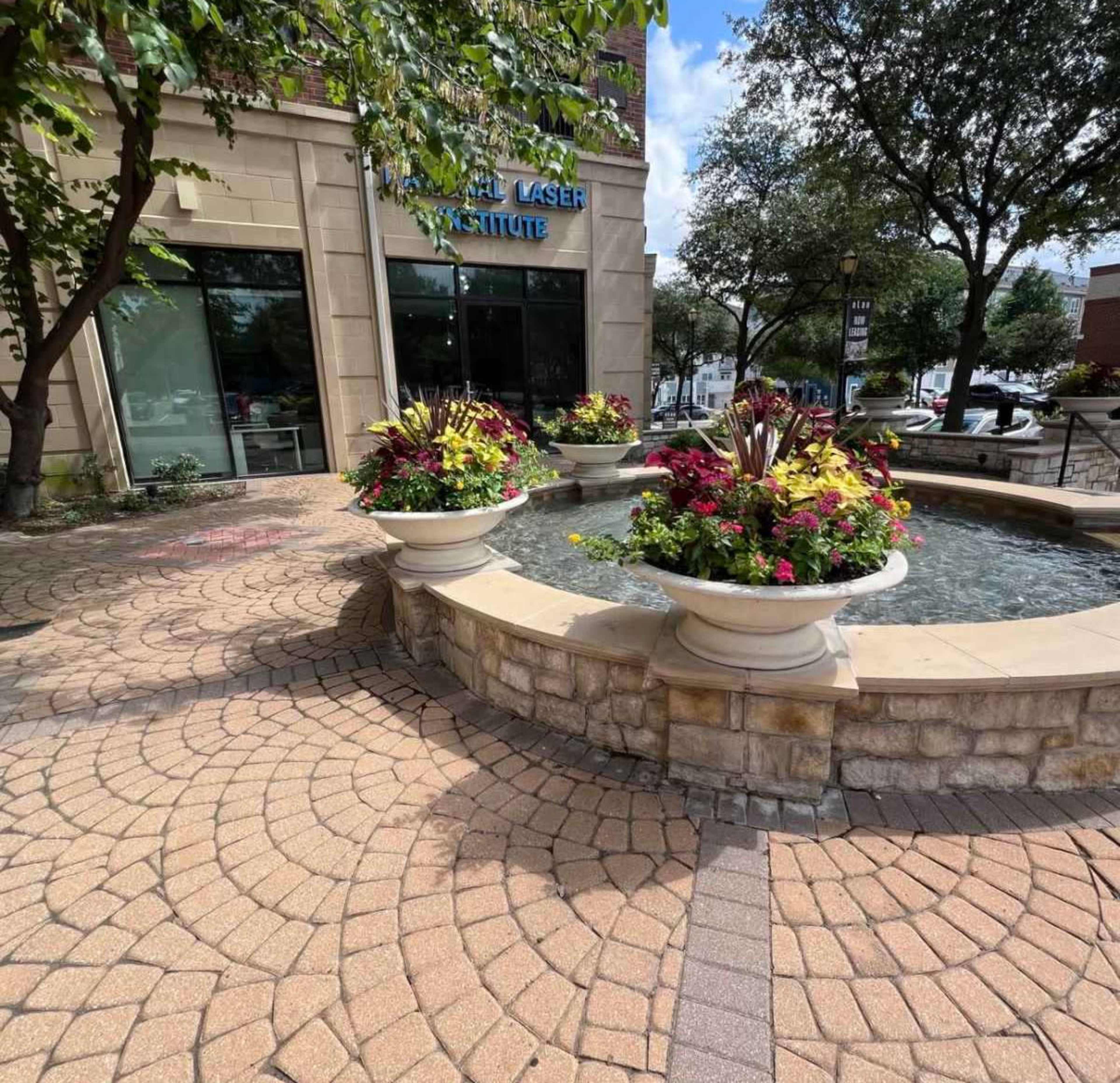 A circular stone fountain surrounded by colorful flower arrangements is situated outside a building labeled "Laser Institute."