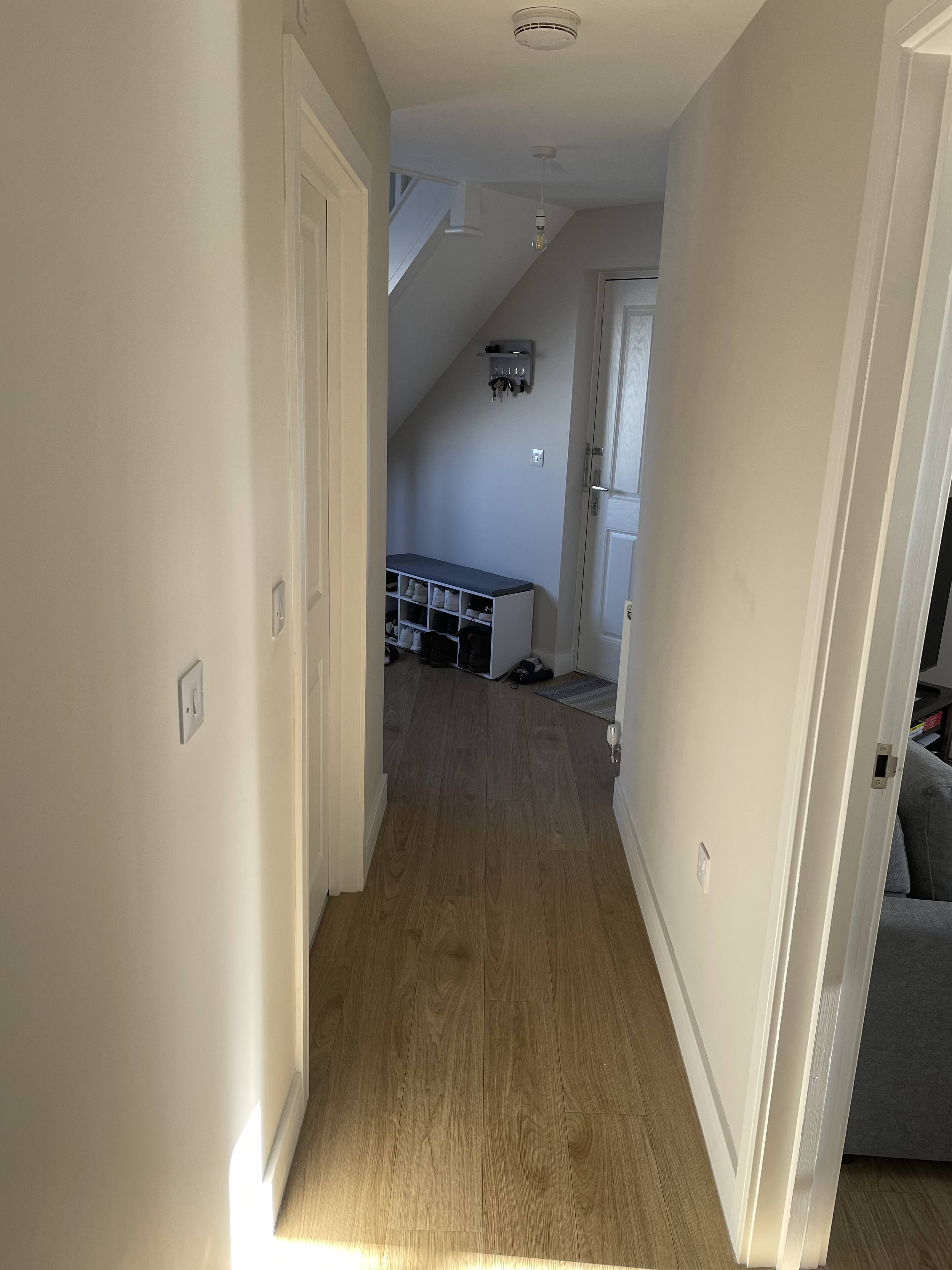 The image shows a narrow hallway with light-colored walls and wooden flooring, leading to a door at the end.