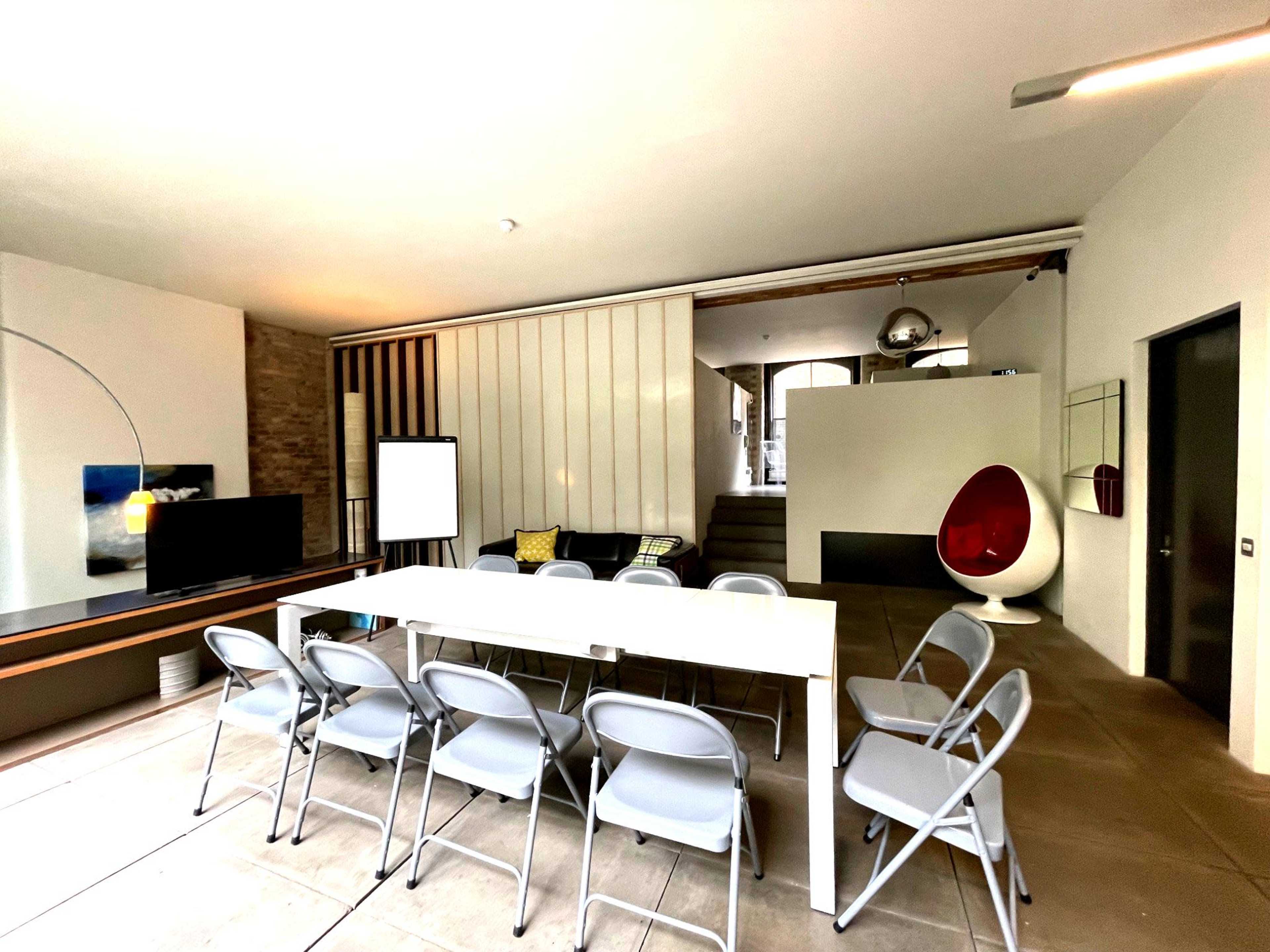 The image shows a modern, open-concept meeting space featuring a long white table with folding chairs, a cozy lounge area with a television, and a distinctive red chair against a light-colored background.