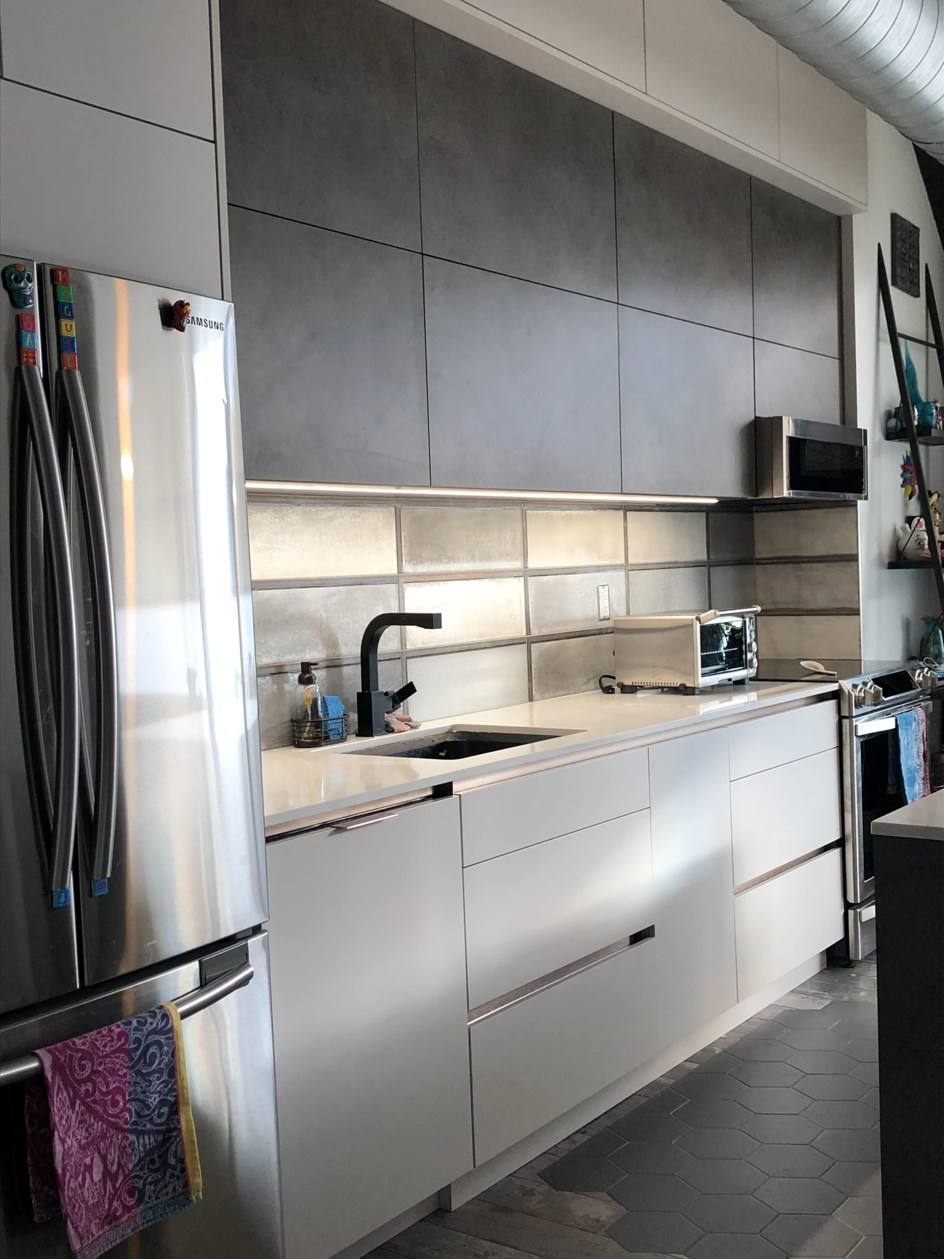 The image shows a modern kitchen with sleek cabinets, a stainless steel refrigerator, and a minimalist countertop design featuring a sink and a microwave.