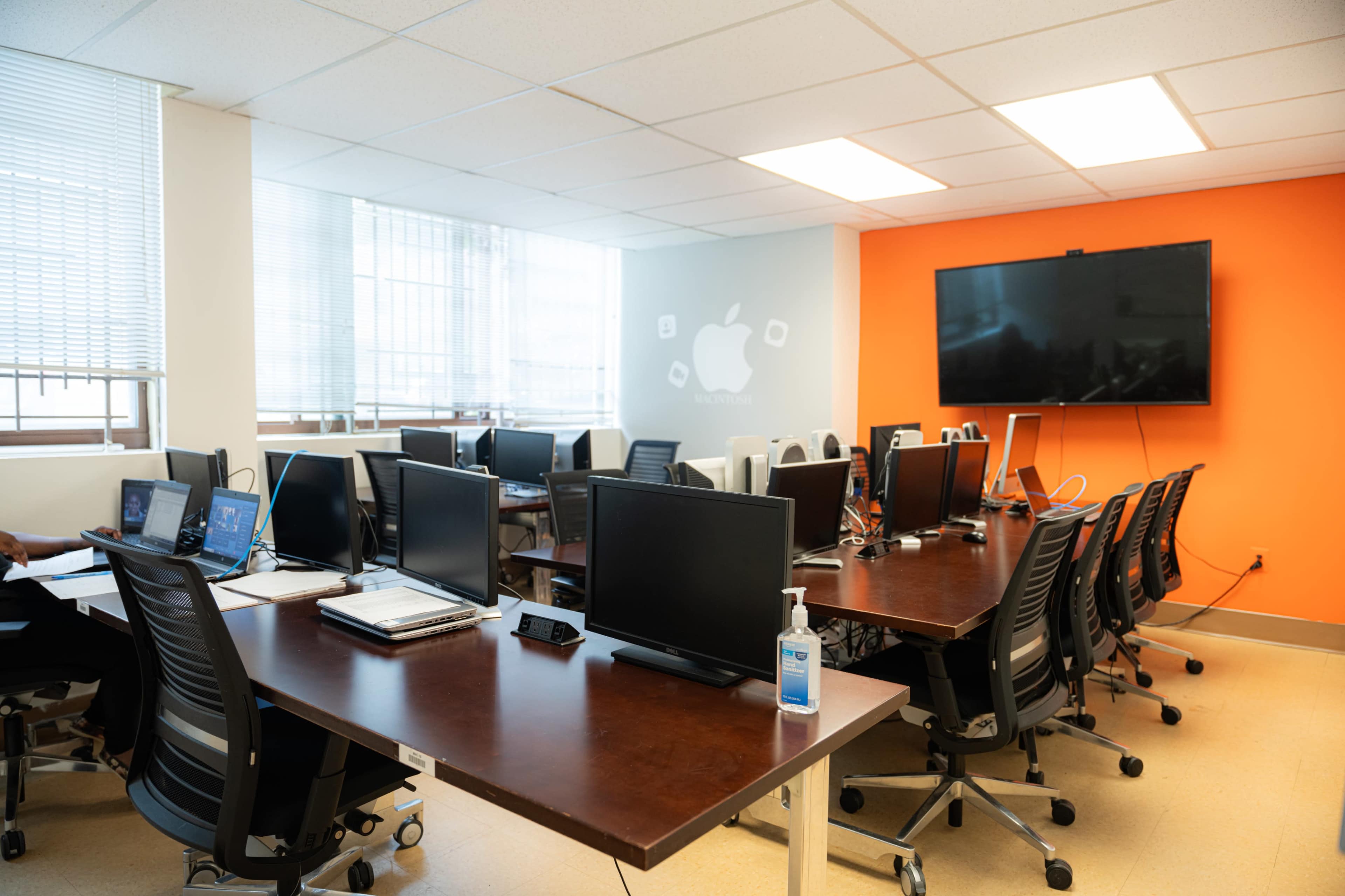 A modern office space features multiple desks with computers and ergonomic chairs, large windows, and a television mounted on an orange wall.