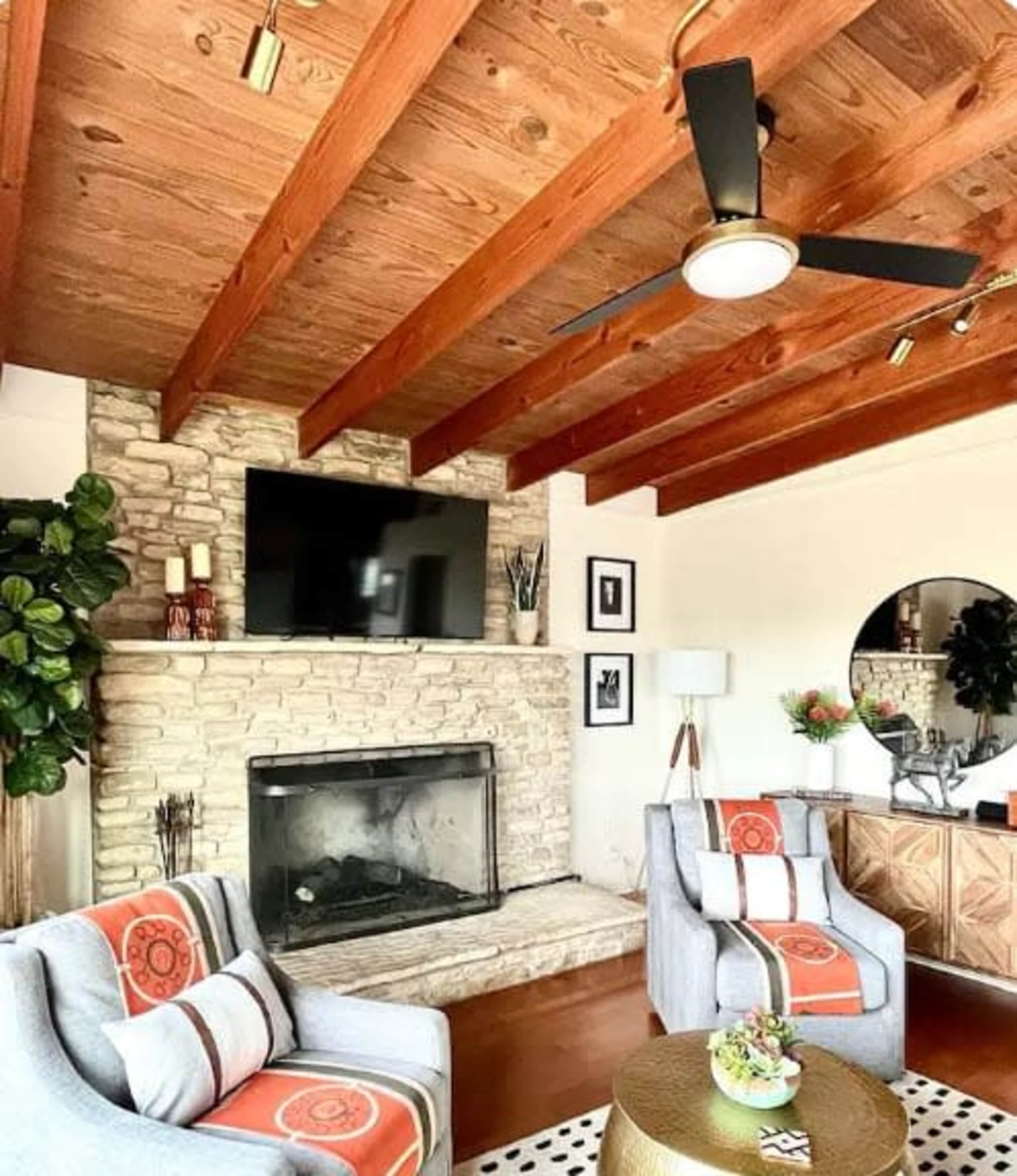 The image shows a living room featuring a stone fireplace, wooden beams, a ceiling fan, two armchairs with throw blankets, and a circular mirror above a wooden side table.
