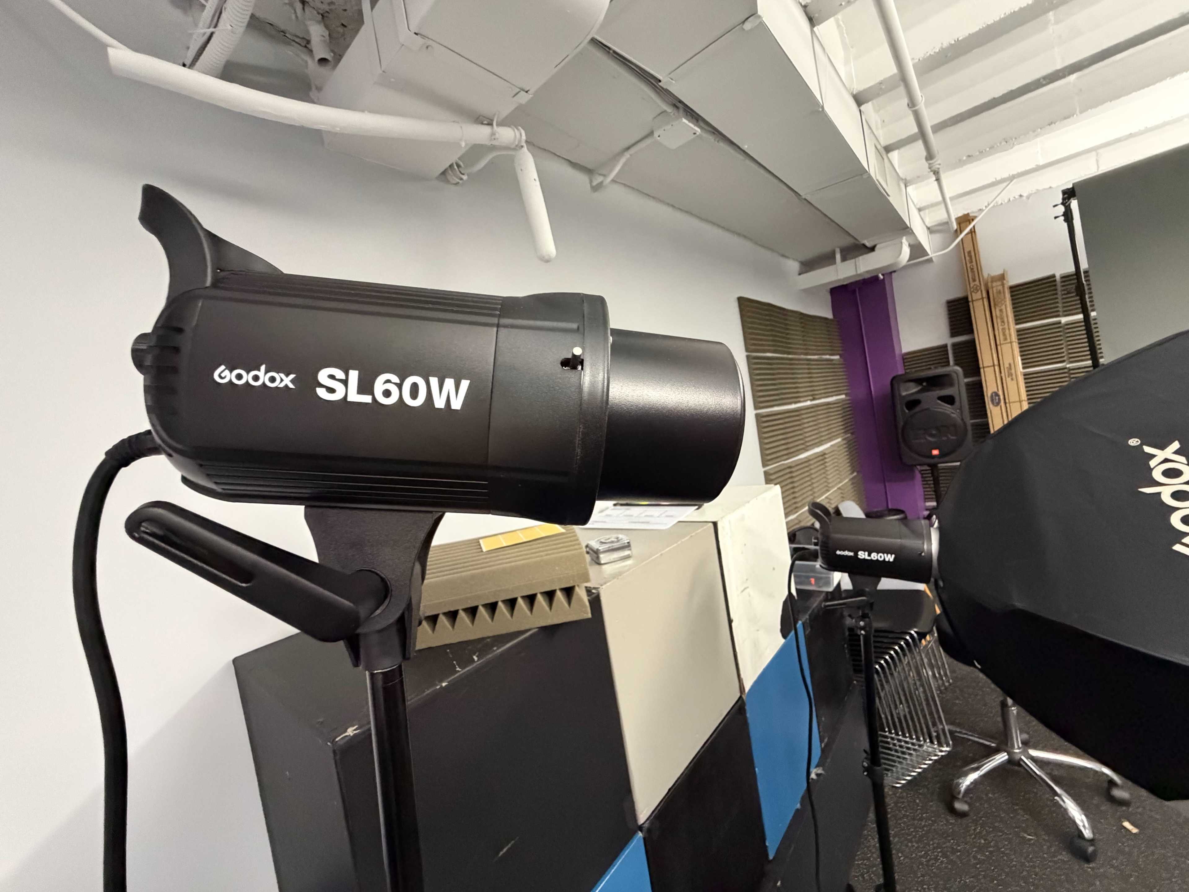 A Godox SL60W studio light is positioned on a stand in a photography studio, surrounded by various backdrops and equipment.