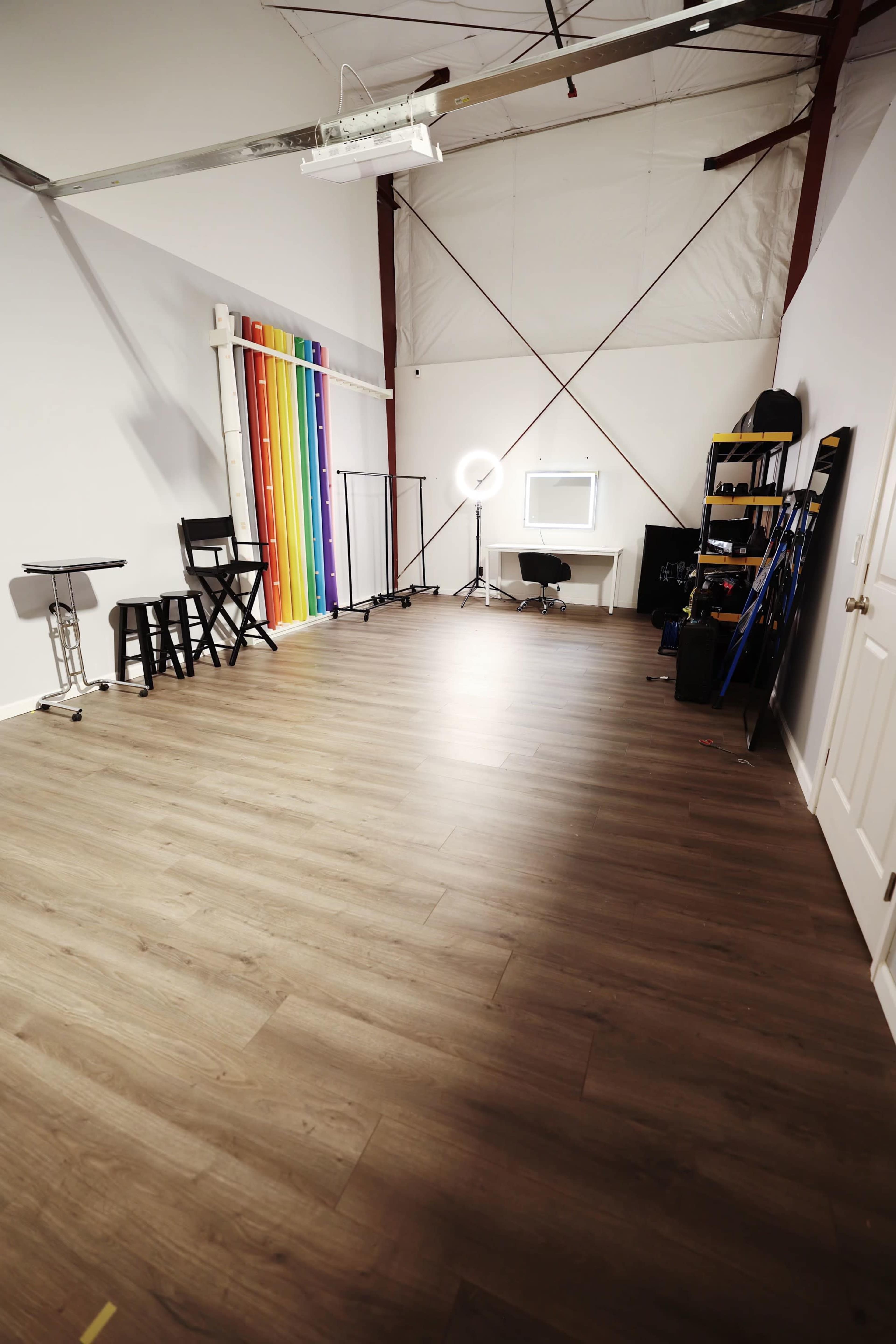 A spacious studio space with light wood flooring, a rainbow backdrop, a ring light, and various furniture items arranged along the walls.
