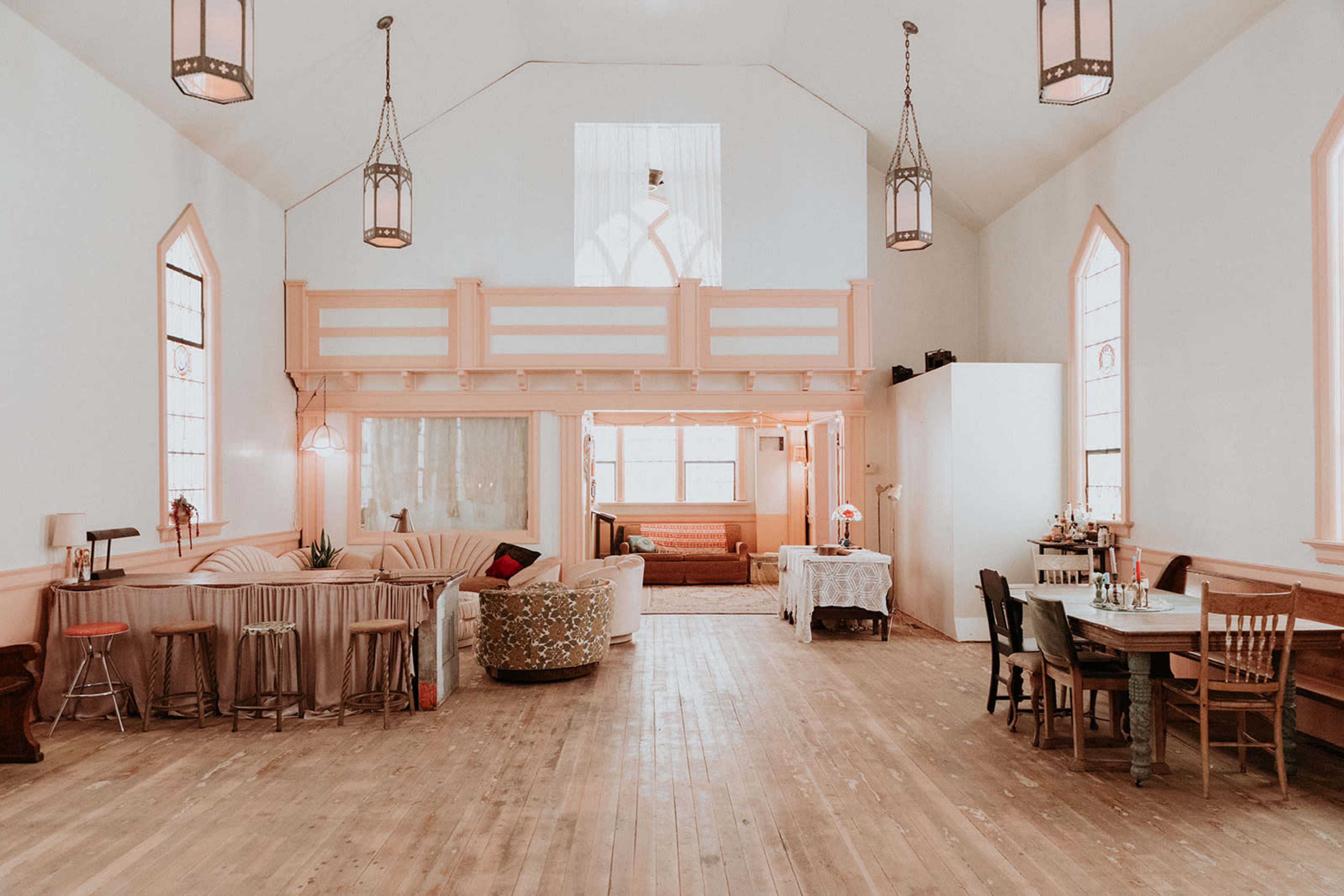 A spacious, light-filled room features wood flooring, tall windows, and a mix of seating areas, including a lounge and dining space.