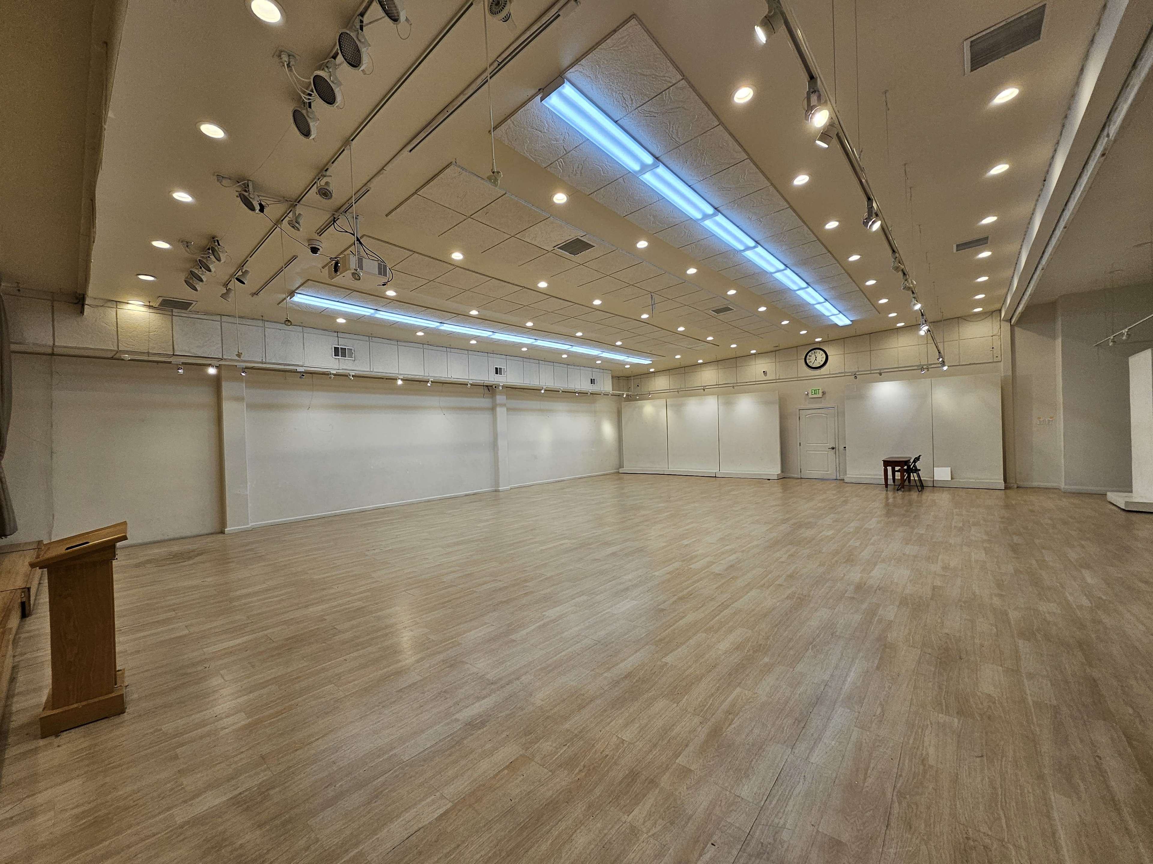 A spacious, well-lit room with wooden flooring and simple white walls, featuring a small table at one end.
