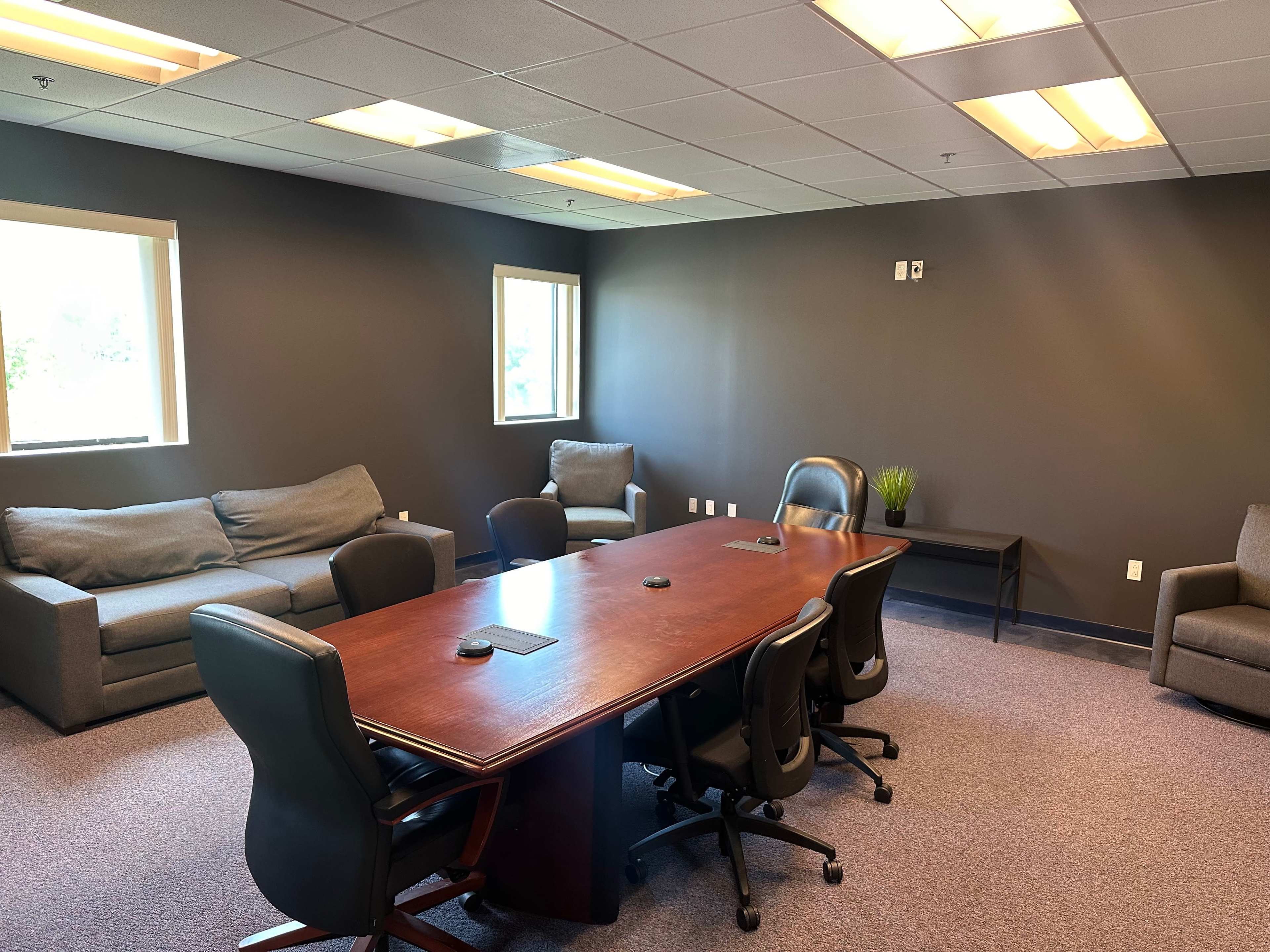 A conference room with a large wooden table surrounded by chairs, flanked by couches and a small side table against a dark wall.