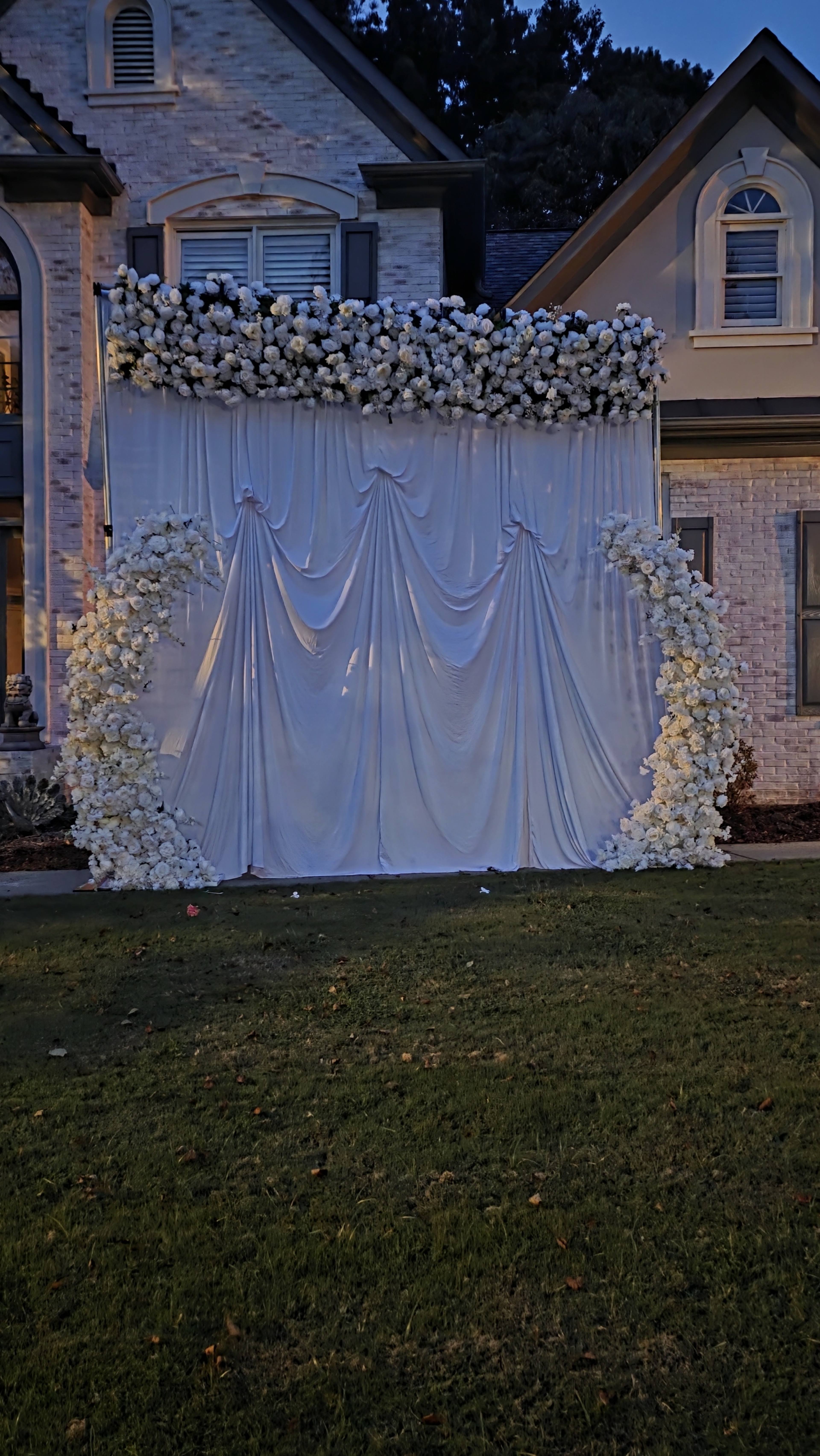 A flower-decorated backdrop with white fabric draping is set up in front of a house.