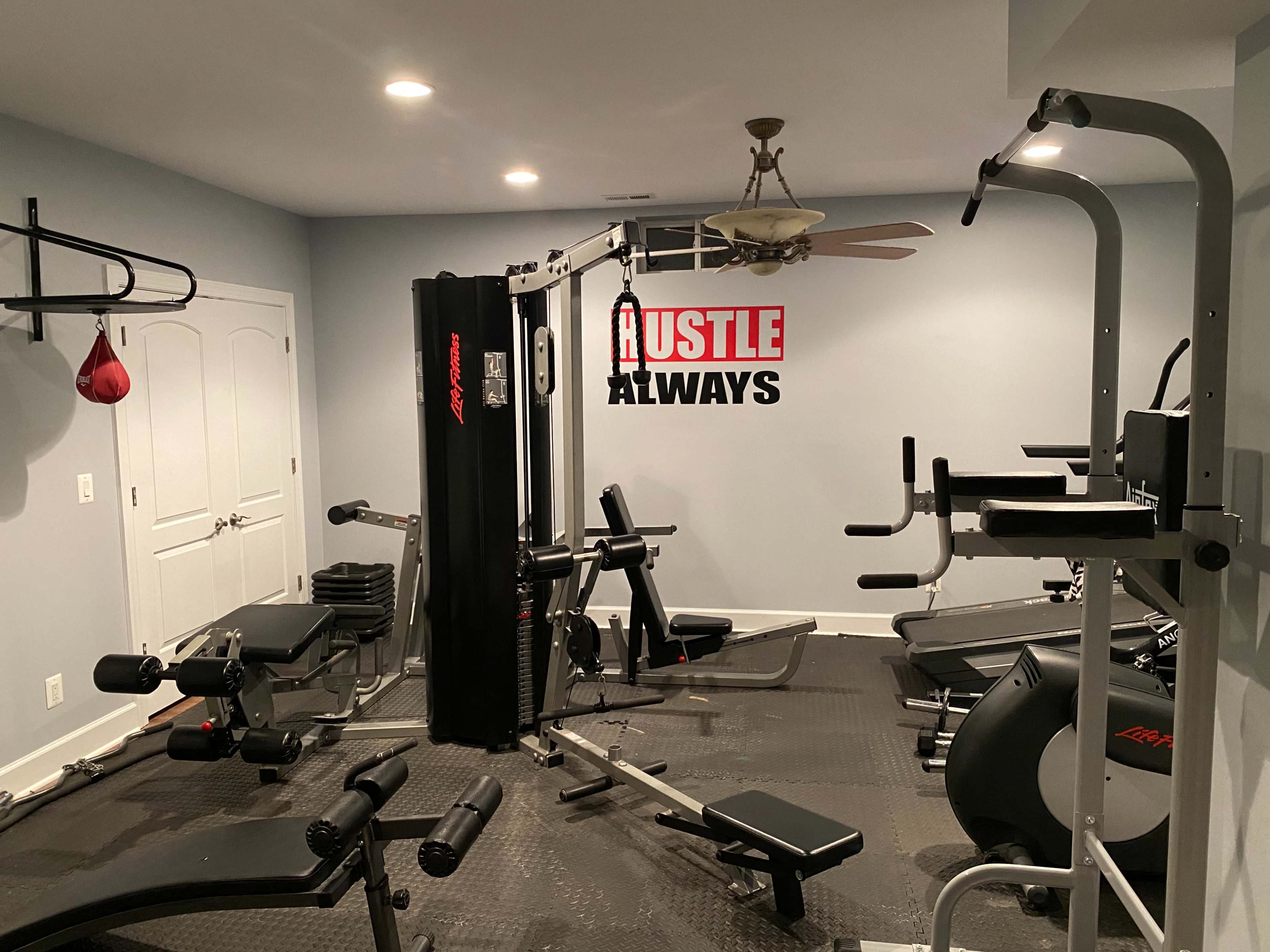 A home gym equipped with various workout machines, weights, and exercise benches, along with a wall featuring the phrase "HUSTLE ALWAYS."