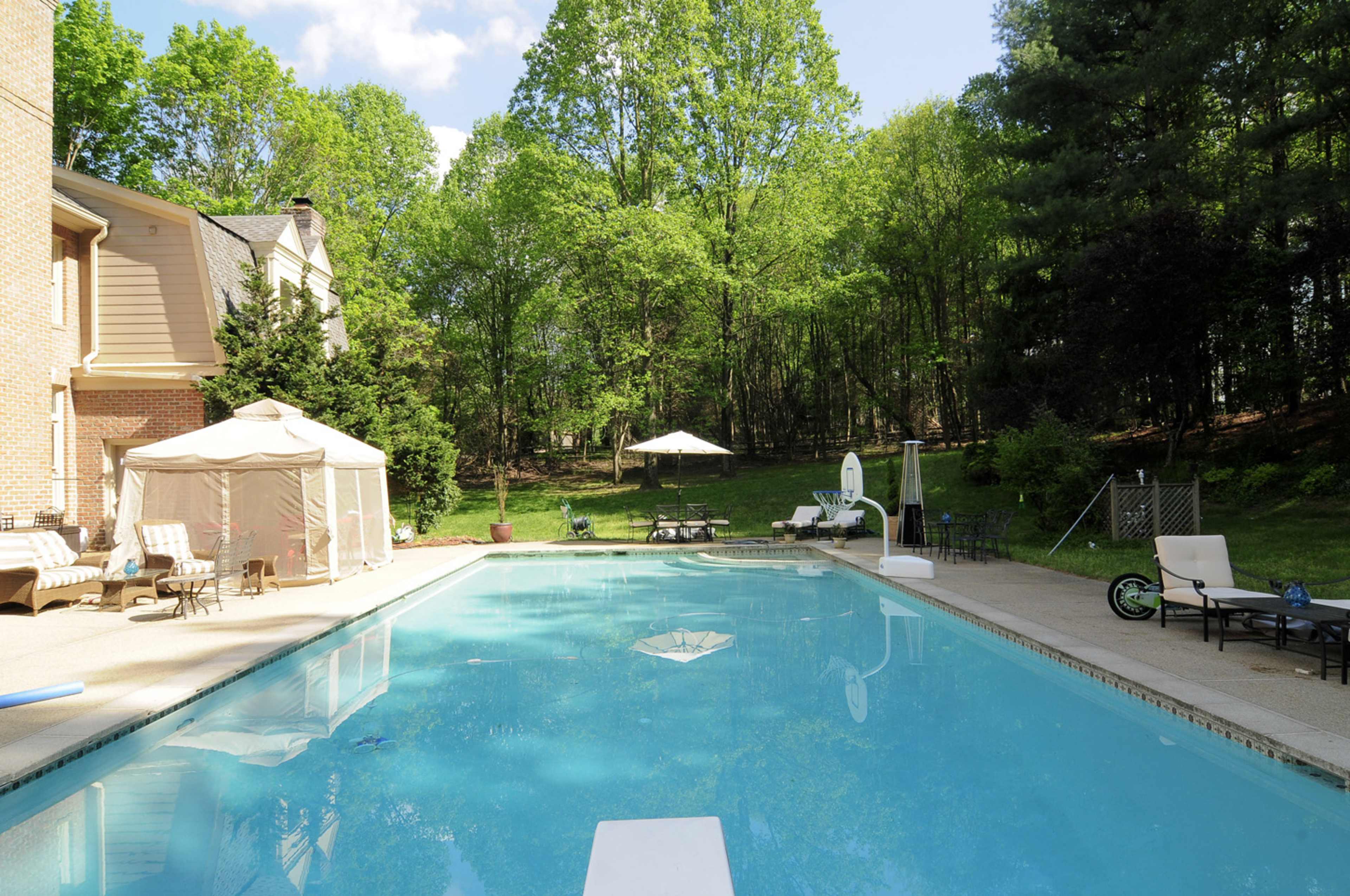 A clear swimming pool surrounded by a deck, lounge chairs, and a gazebo in a wooded backyard.