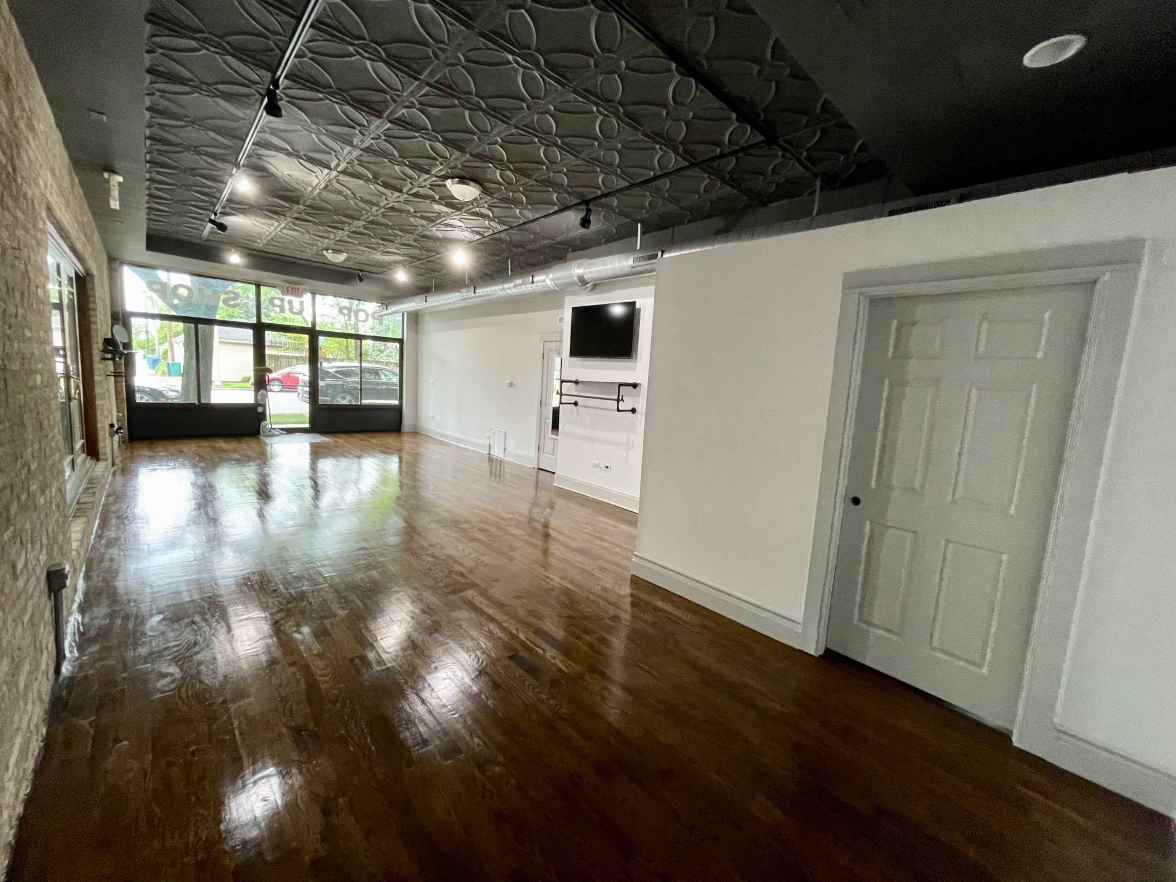 The image shows an empty room with hardwood floors, a textured ceiling, a wall-mounted screen, and a door on the right side.