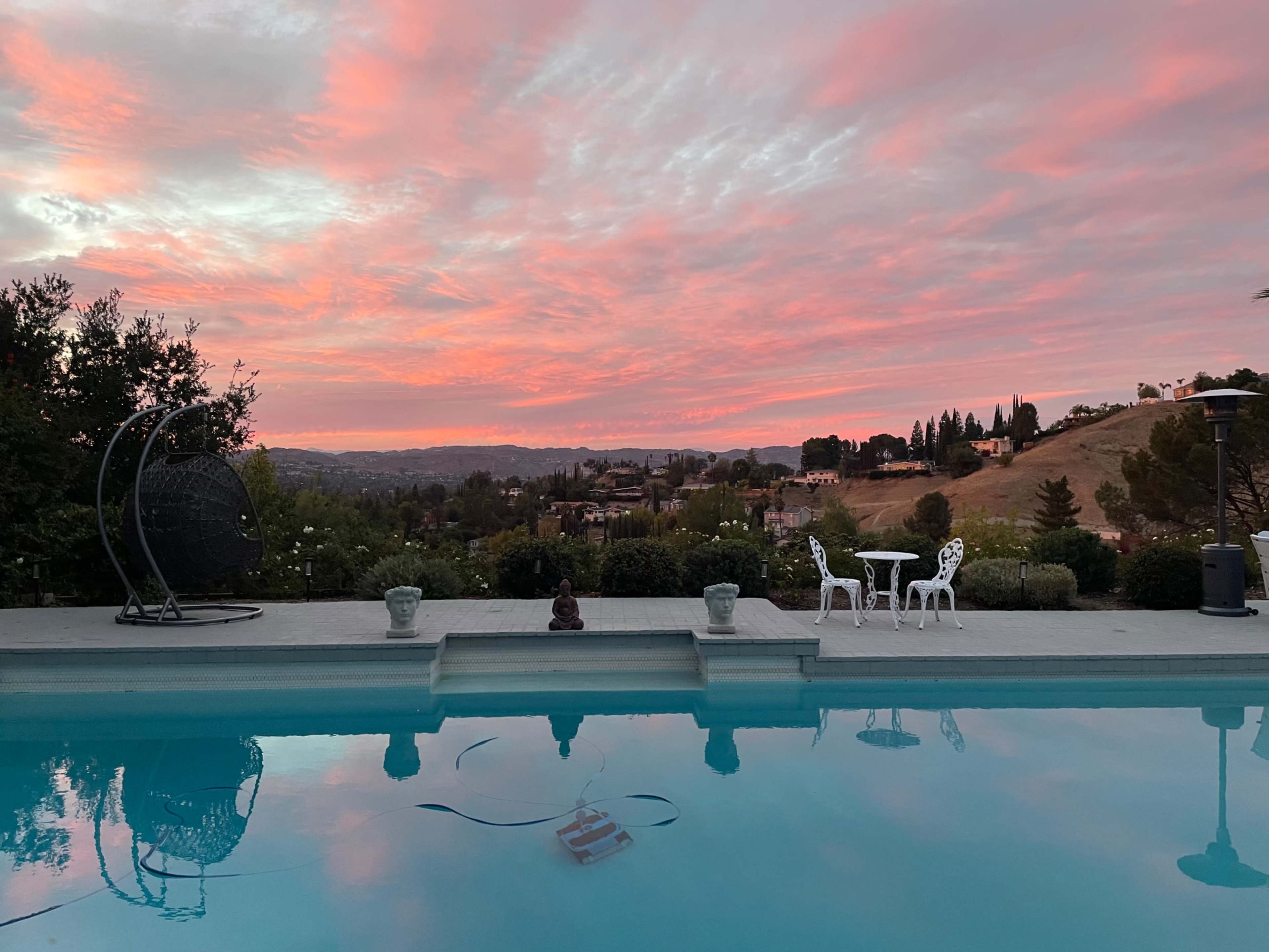 The scene features a swimming pool reflecting a colorful sunset sky, with a hillside view and two white chairs set near the water's edge.