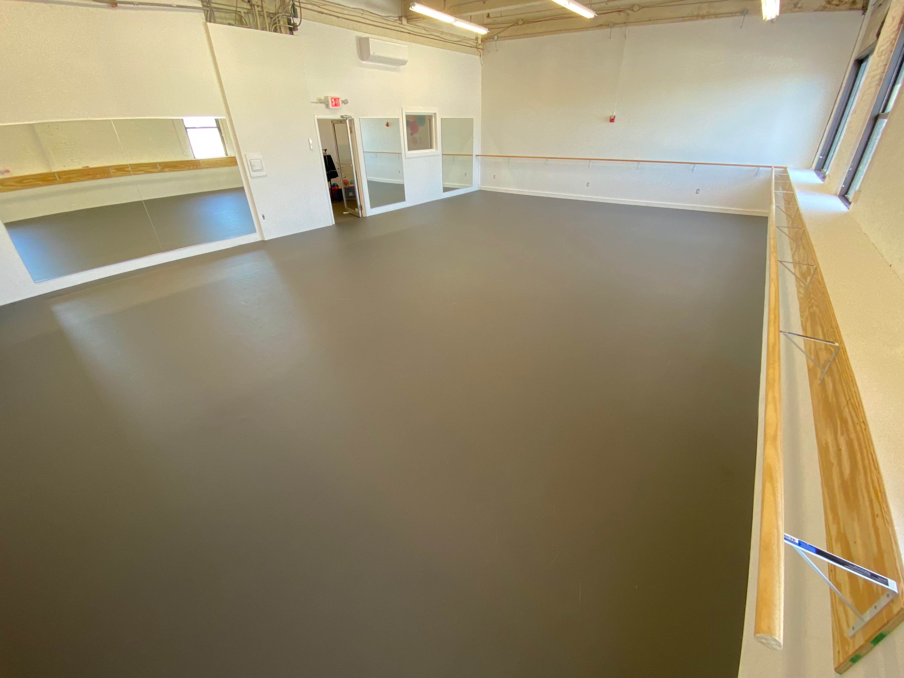A spacious dance studio with a smooth gray floor, large mirrors on one wall, and a ballet barre running along another wall.