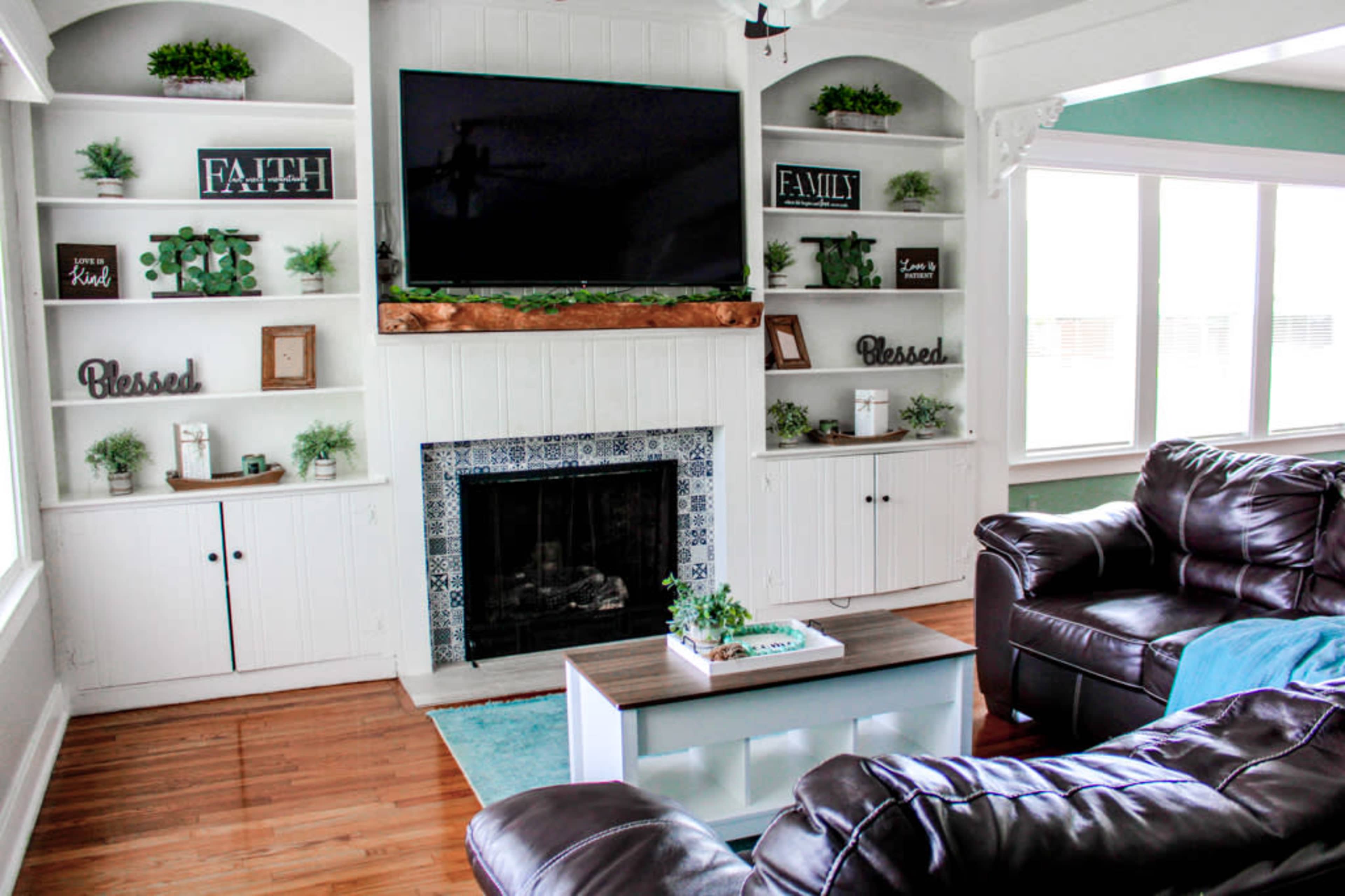 A living room with a fireplace, a large television mounted above it, and built-in shelves decorated with plants and framed signs, alongside a coffee table and dark leather sofas.