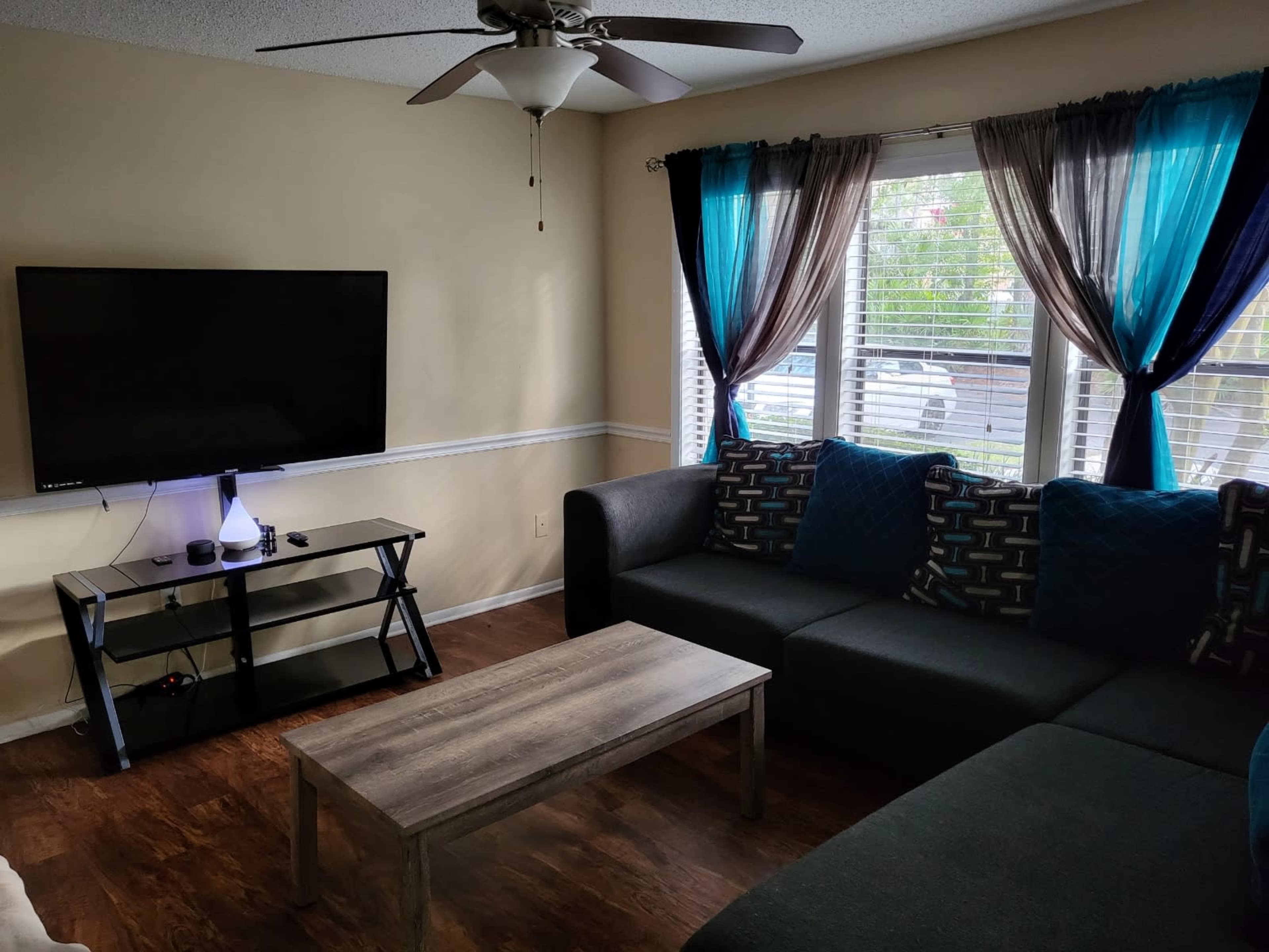 The living room features a dark sectional sofa, a wooden coffee table, a mounted television, and large windows covered with blue and black curtains.