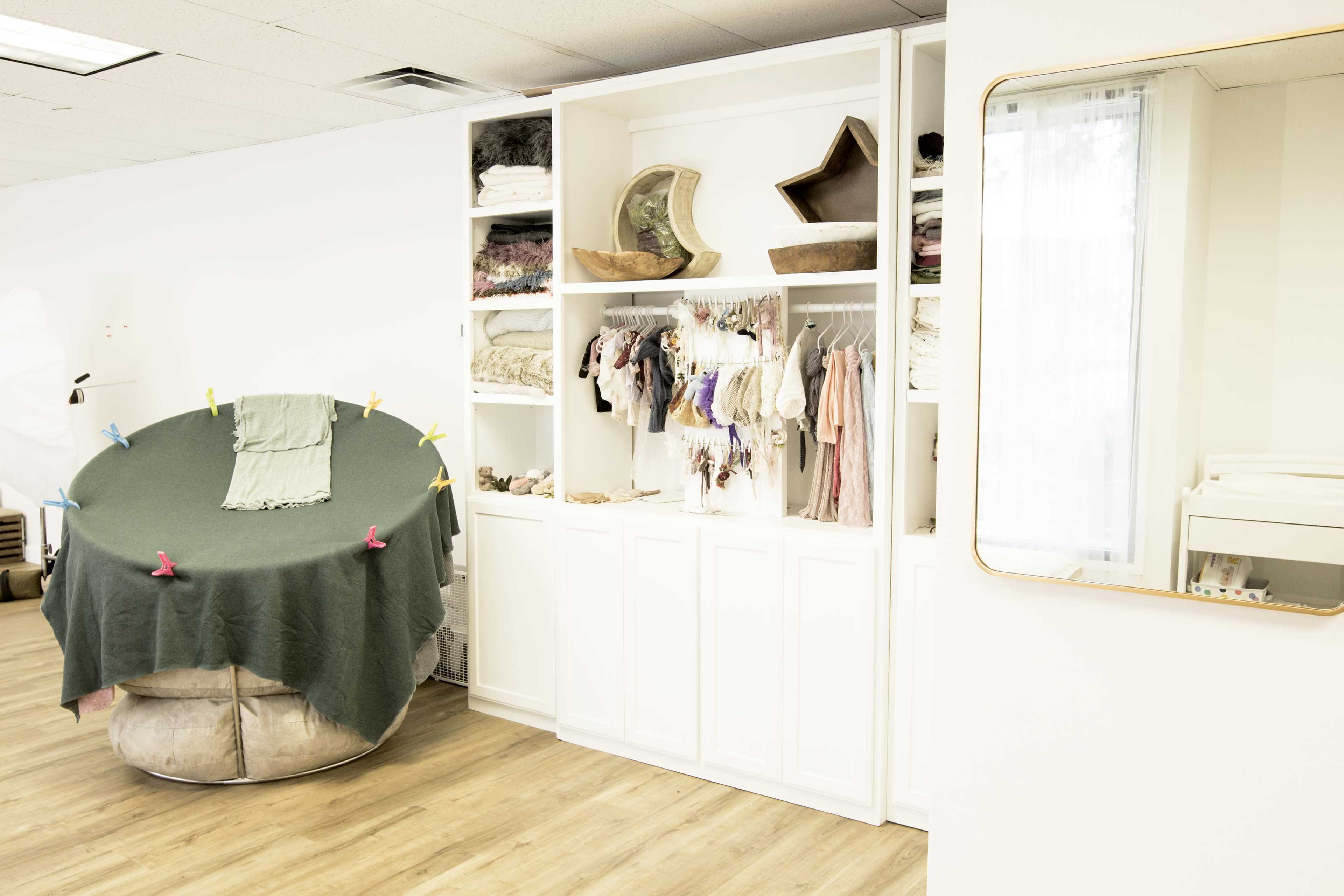 The image shows a brightly lit room featuring a large round seating area covered with a green blanket and a shelving unit filled with neatly arranged fabric items and clothing.