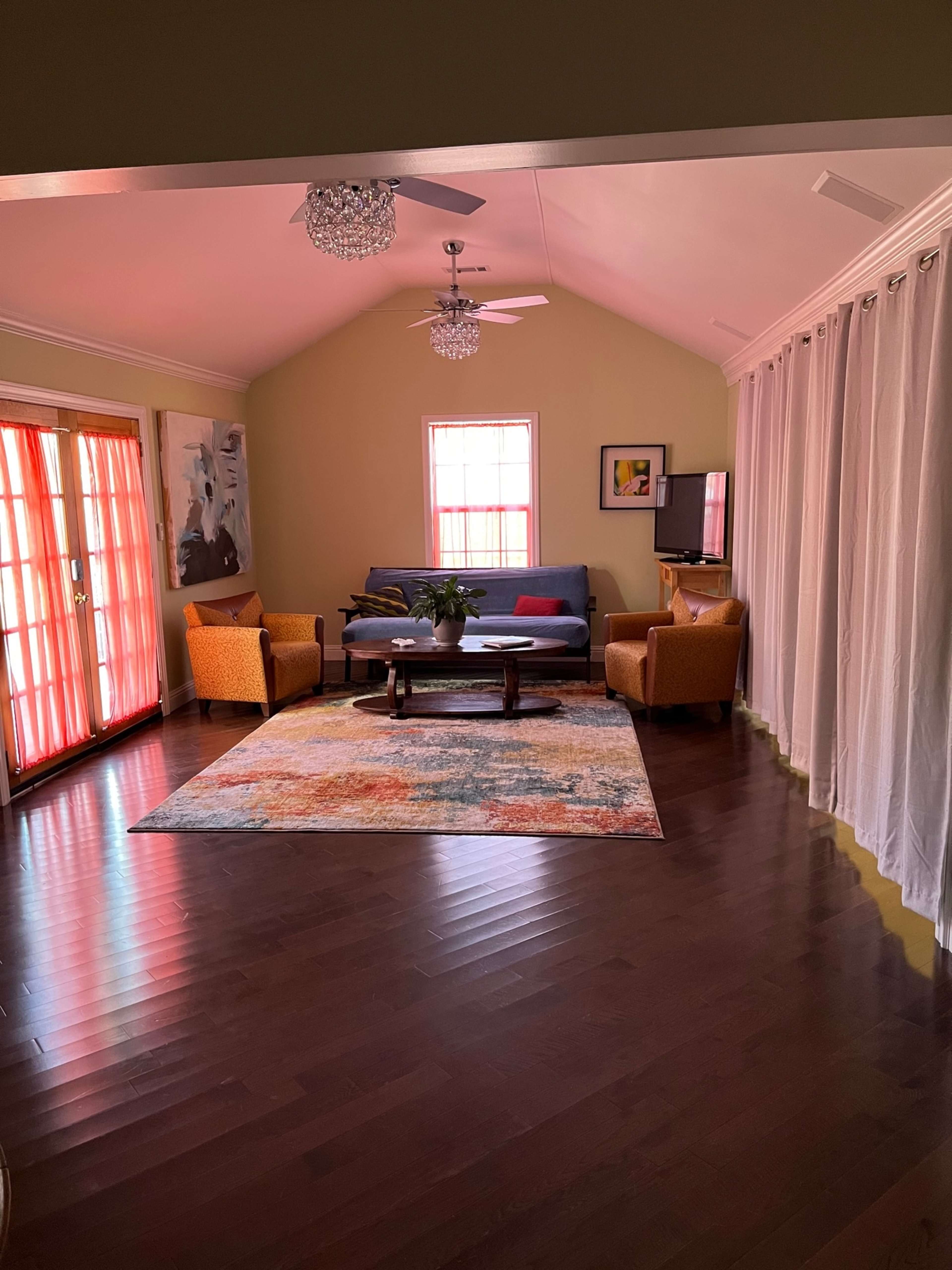 A bright living room with a blue sofa, orange armchairs, and a colorful area rug, illuminated by natural light filtering through sheer curtains and a glass door.