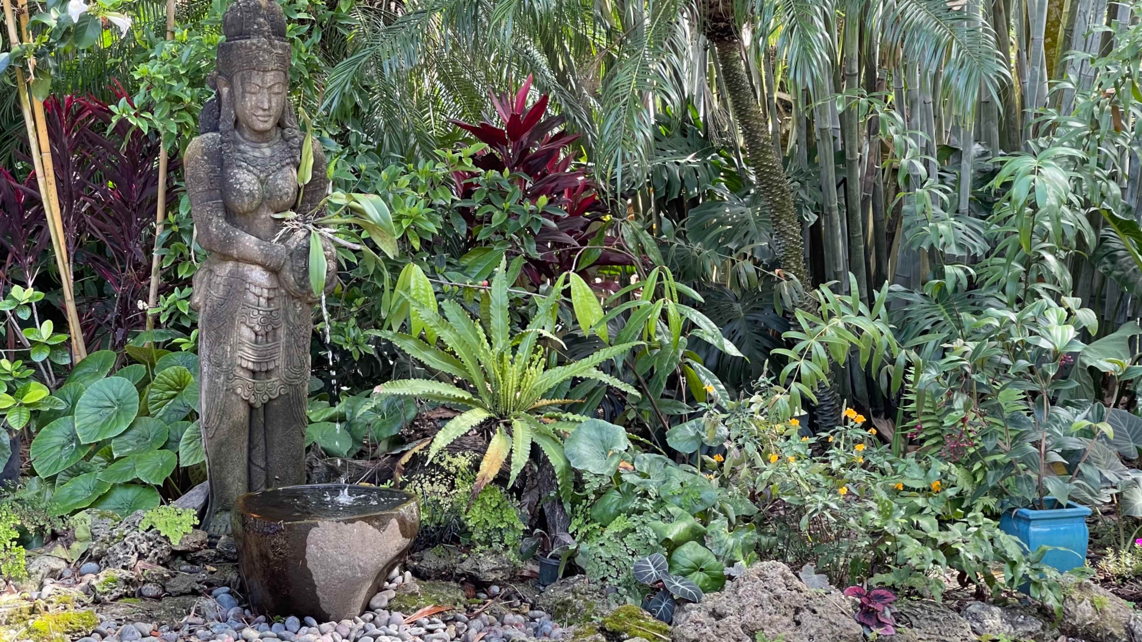 A stone statue of a woman stands beside a round water basin, surrounded by lush tropical plants and flowers.