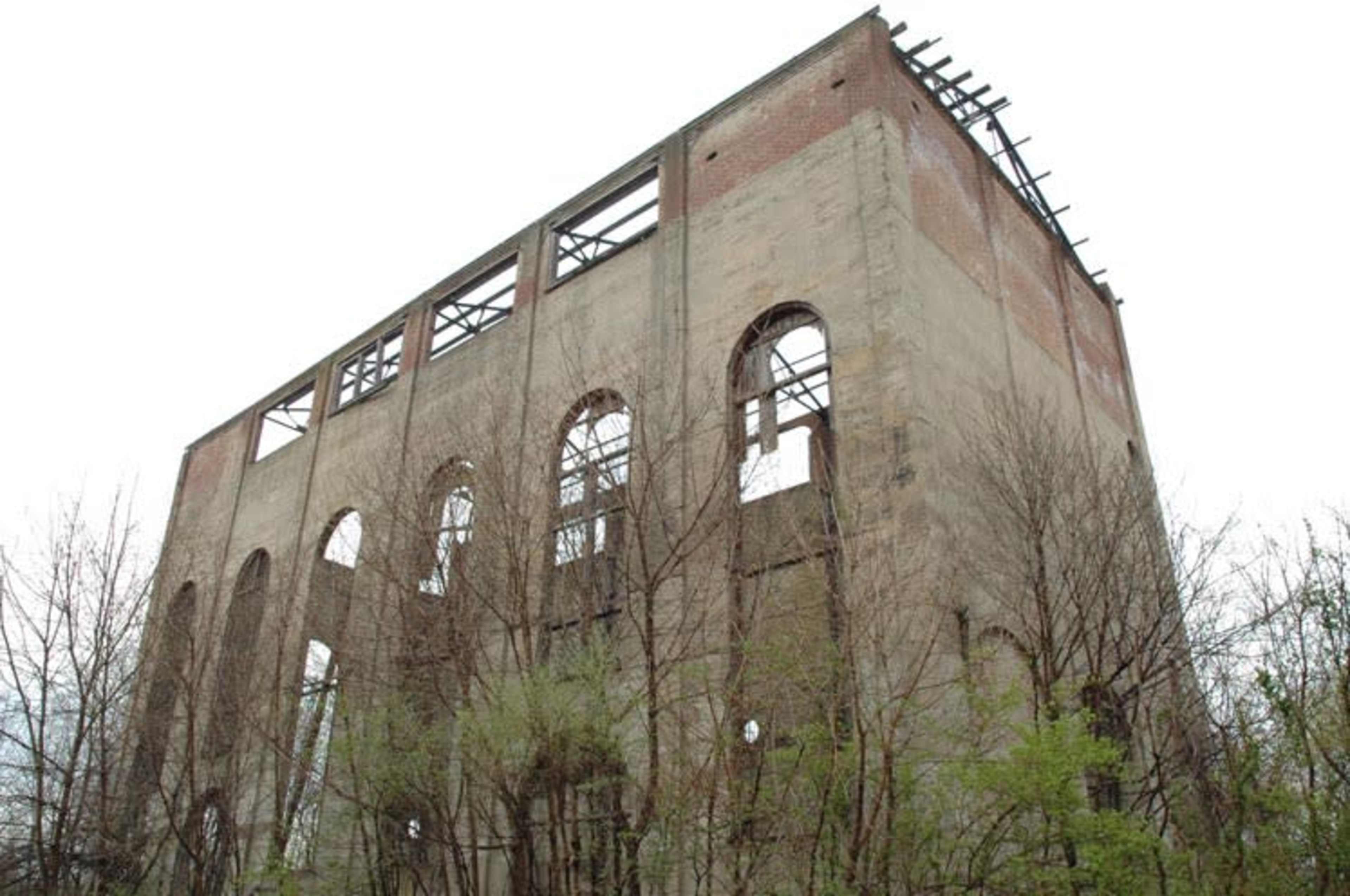 The image shows the remains of a tall, abandoned industrial building with partially collapsed walls and overgrown vegetation surrounding it.