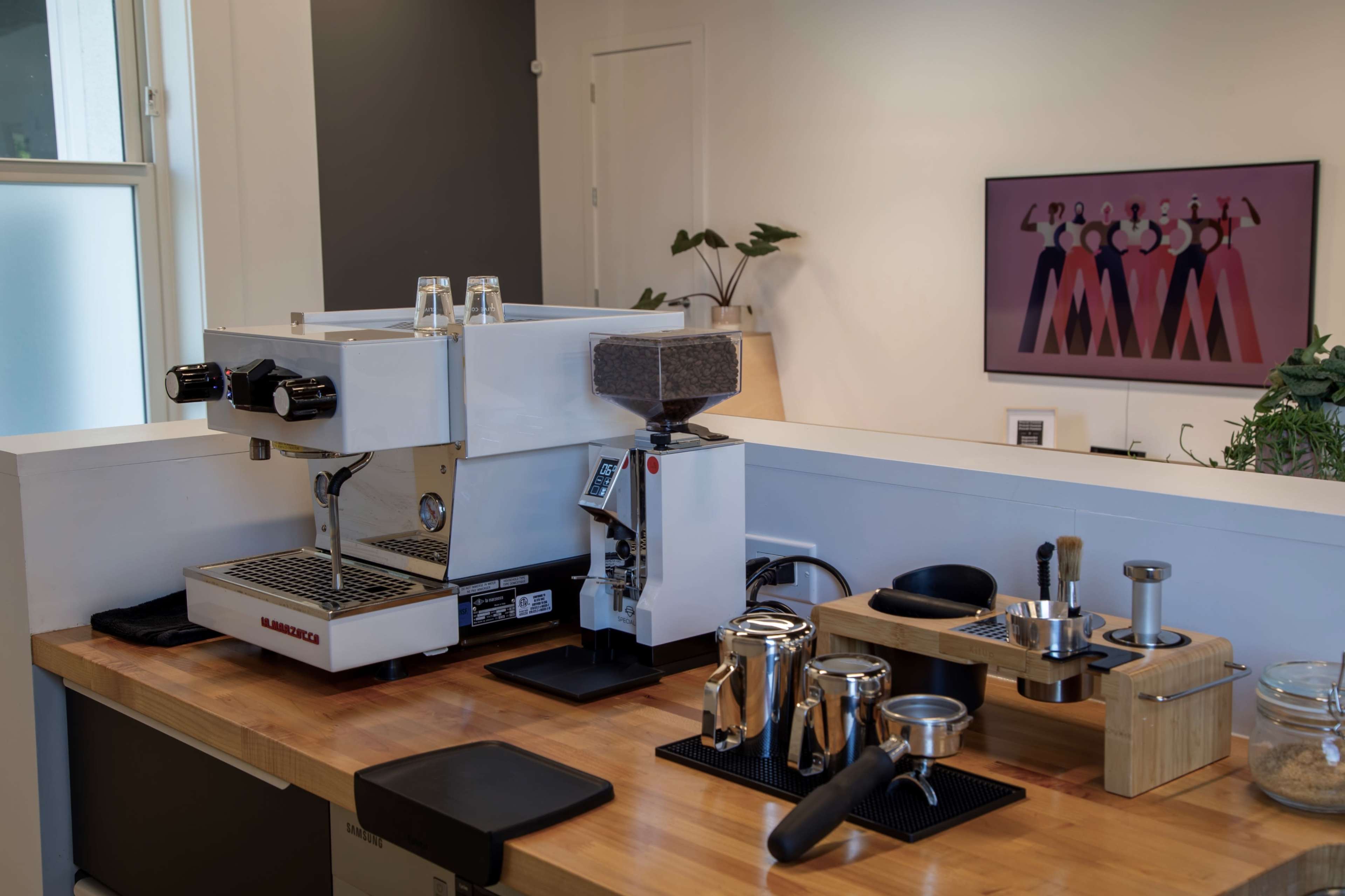 A modern coffee setup featuring an espresso machine, grinder, and various coffee brewing tools on a wooden countertop, with a colorful artwork displayed on the wall behind.