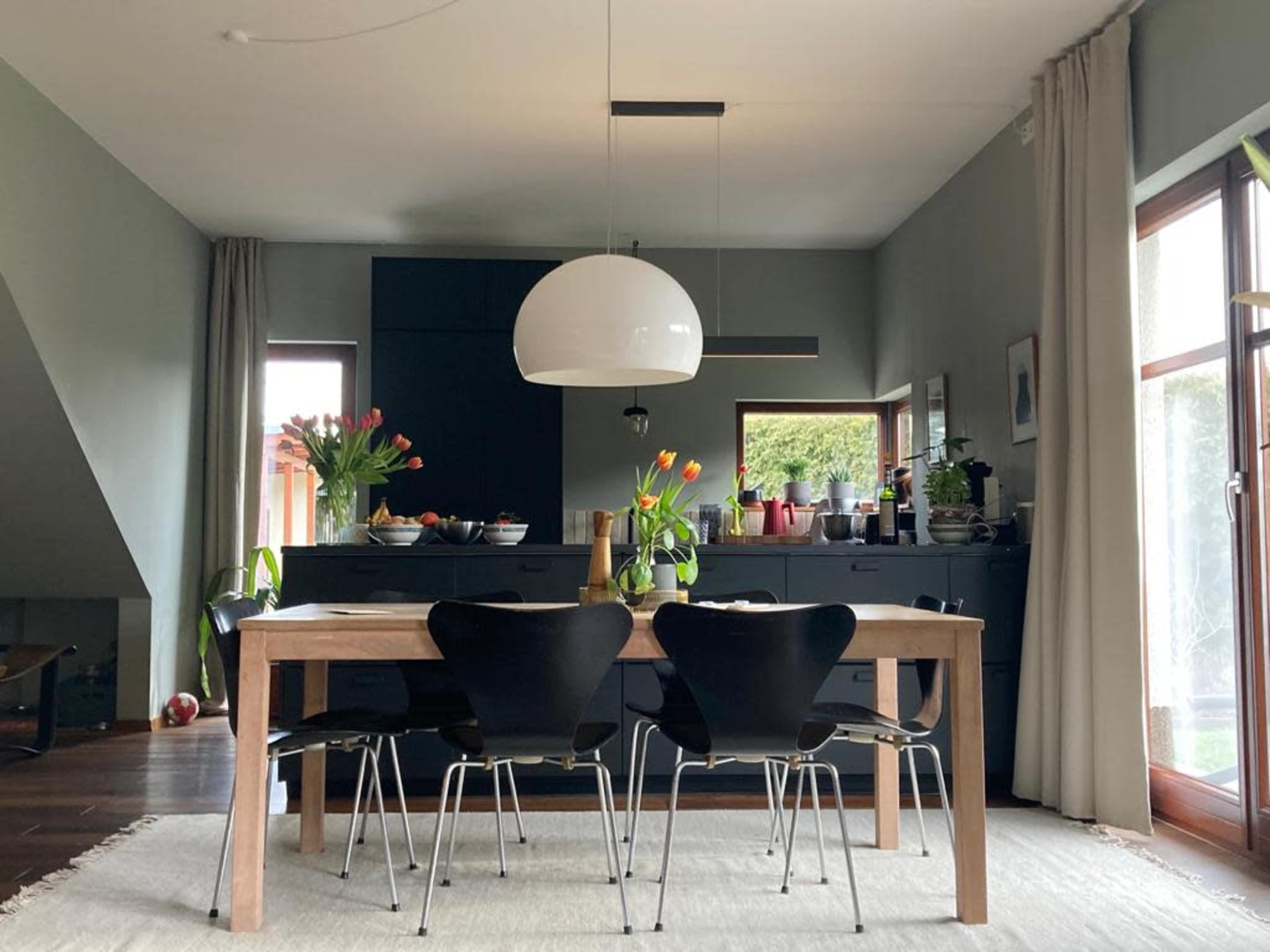 A dining room features a wooden table surrounded by black chairs, with a large pendant light above and a dark cabinetry wall in the background.