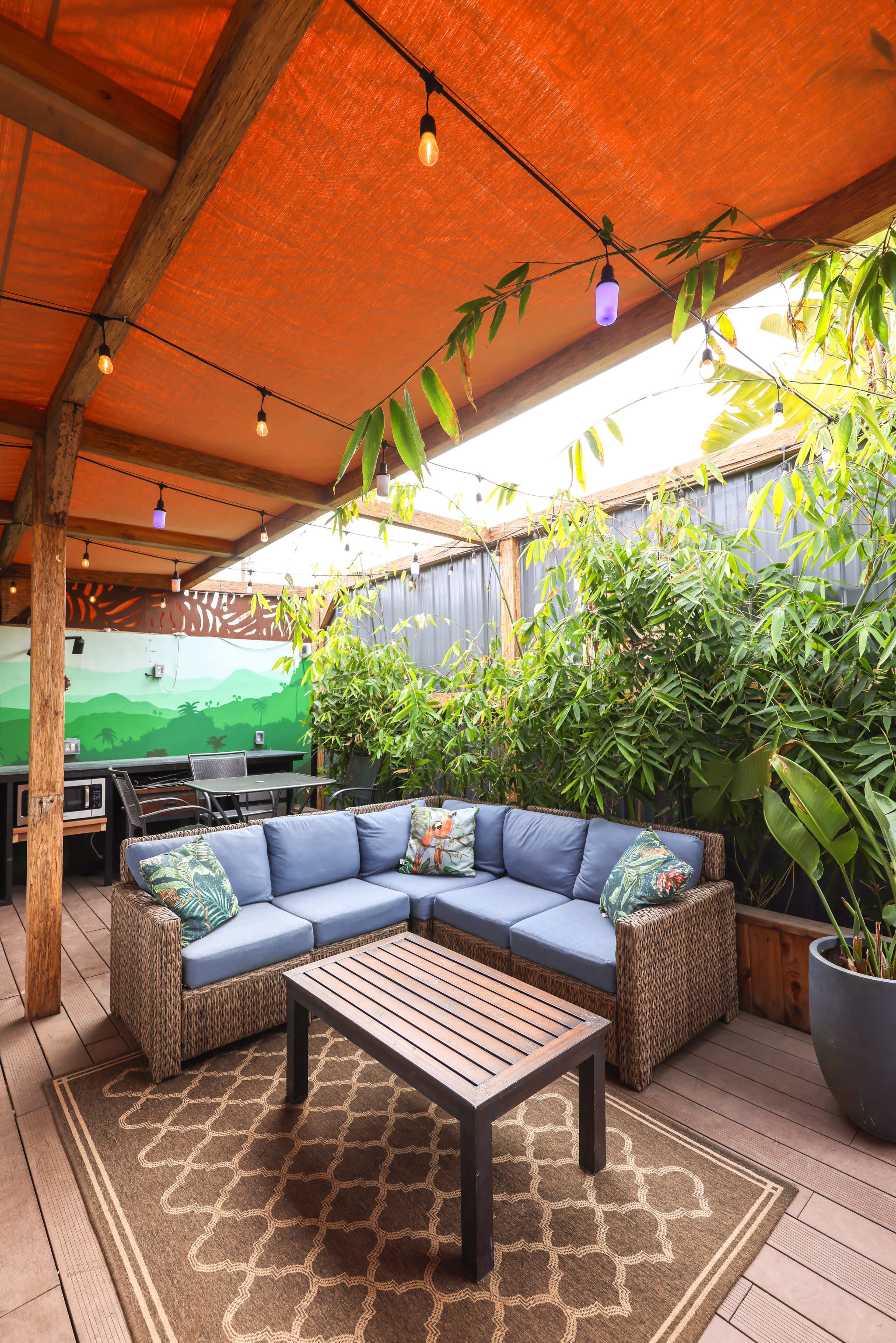 A cozy outdoor patio featuring a sectional sofa, a wooden coffee table, and vibrant greenery underneath a canopy with string lights.