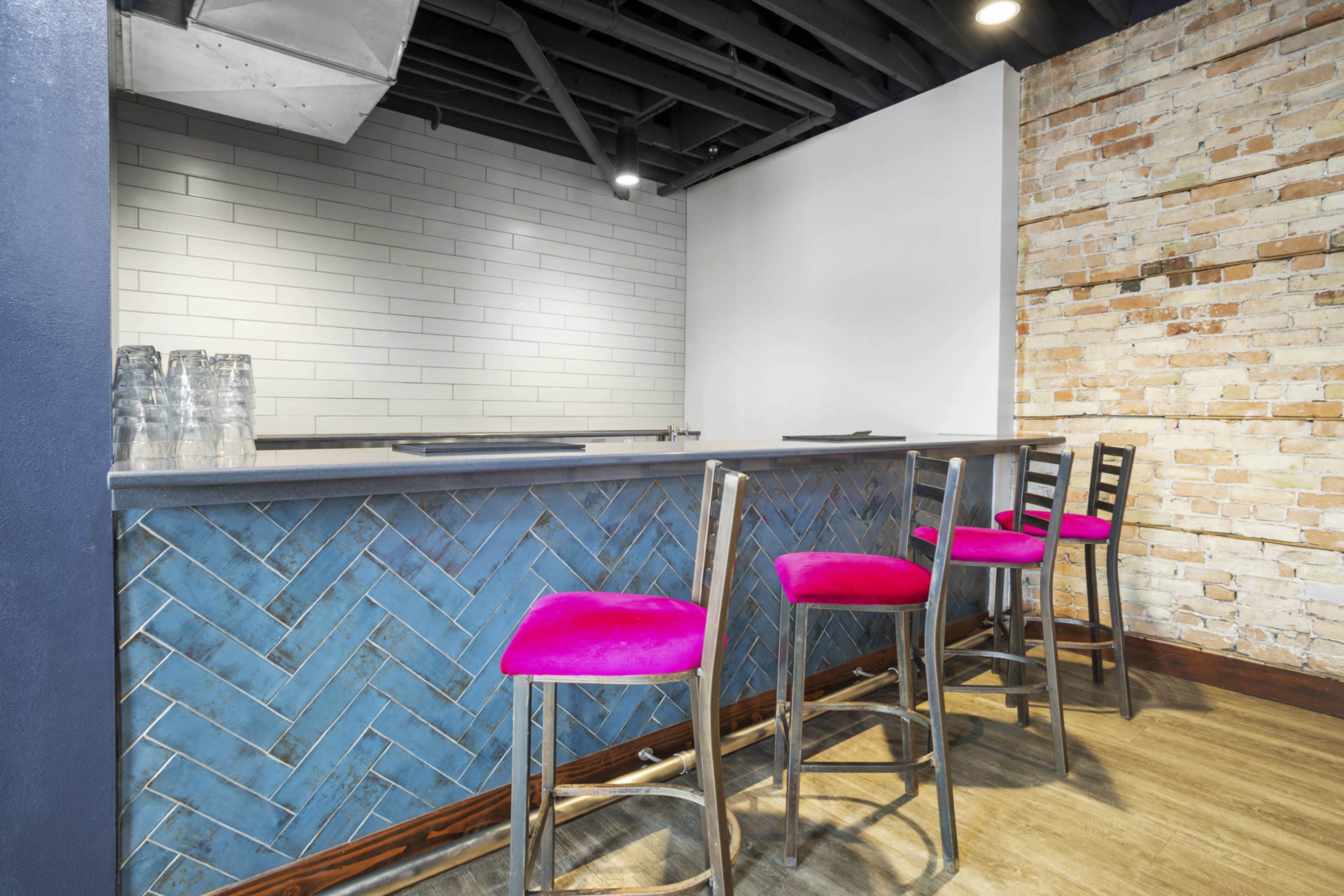 The image shows a modern bar area with a herringbone-patterned blue tile front, a gray countertop, and several high stools with bright pink seat cushions.