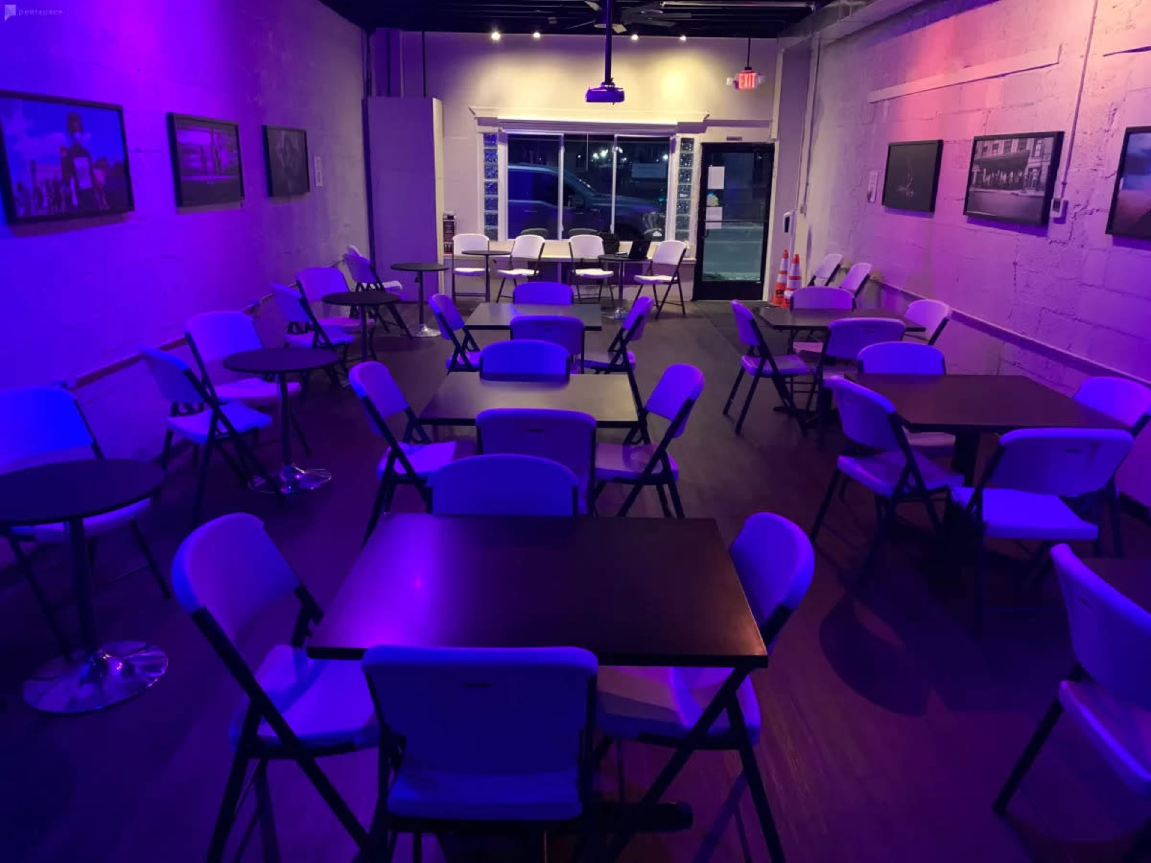 A dimly lit room with several empty folding tables and chairs arranged in a grid pattern, featuring purple lighting and framed photographs on the walls.