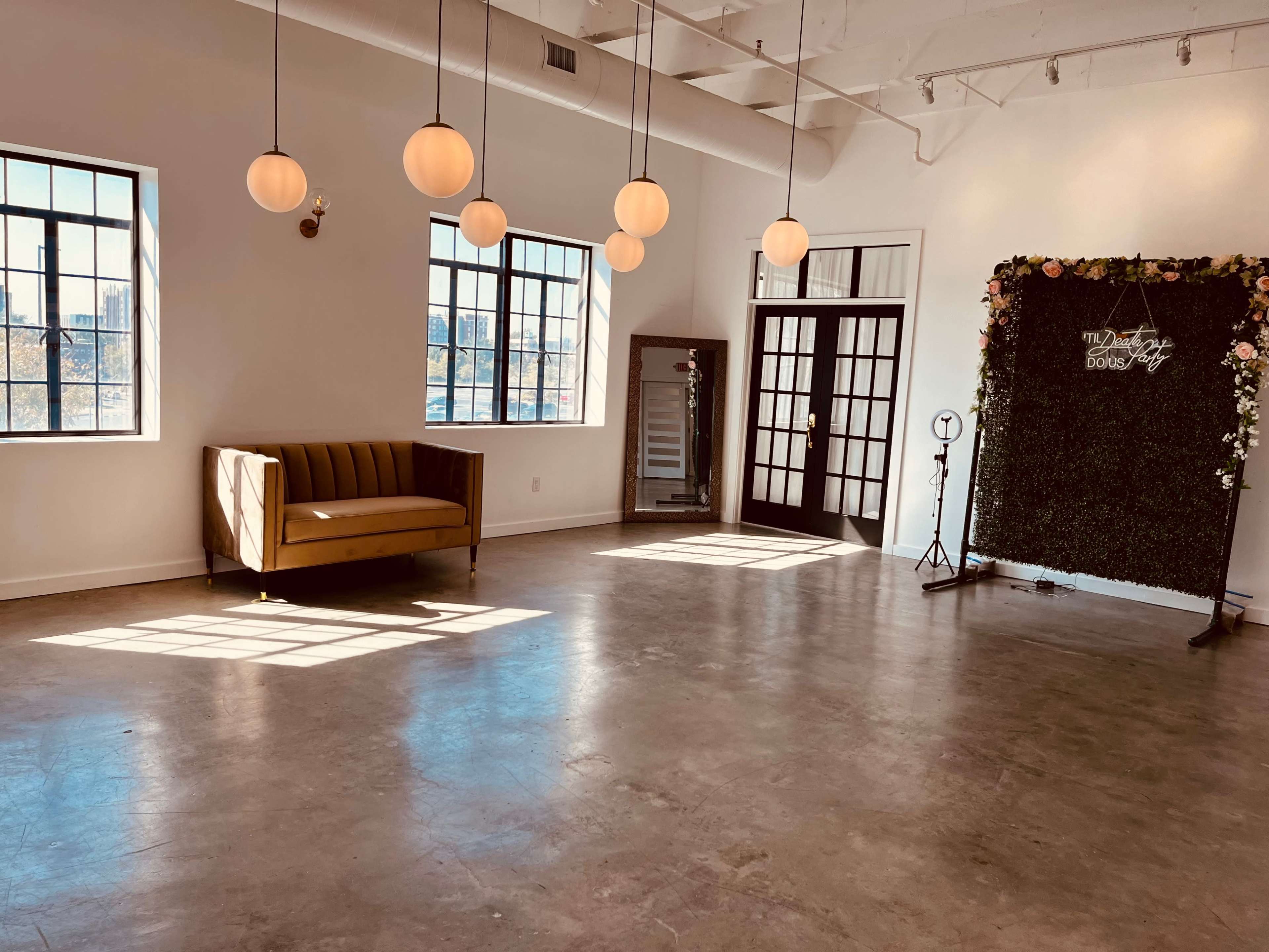 The image shows a bright, spacious room featuring large windows, a vintage-style couch, and a decorative wall with flowers.