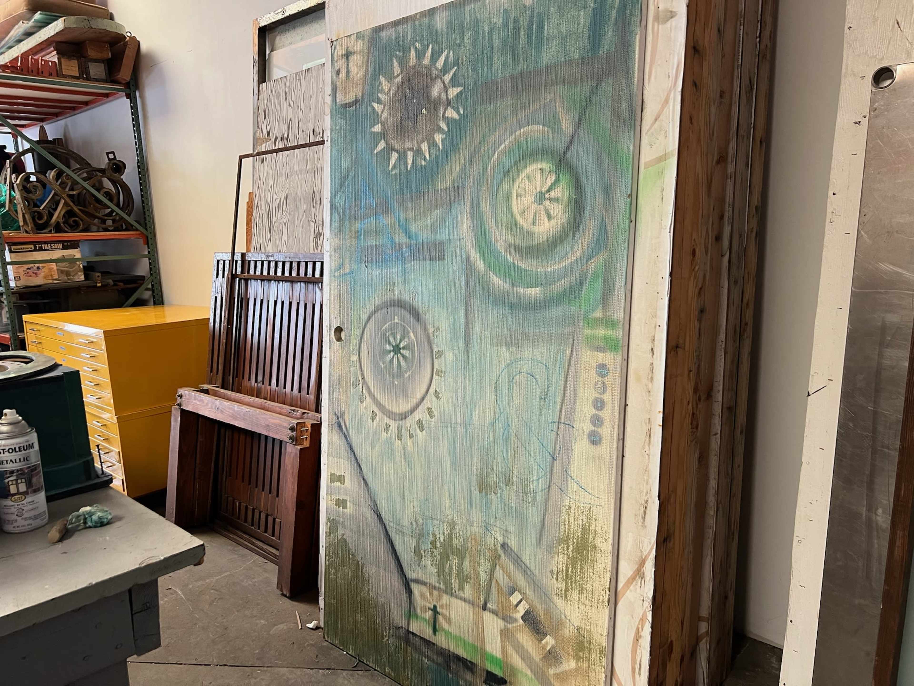The image shows a colorful, abstract painting on a door set against a backdrop of wooden panels and various furniture items in a warehouse or storage area.