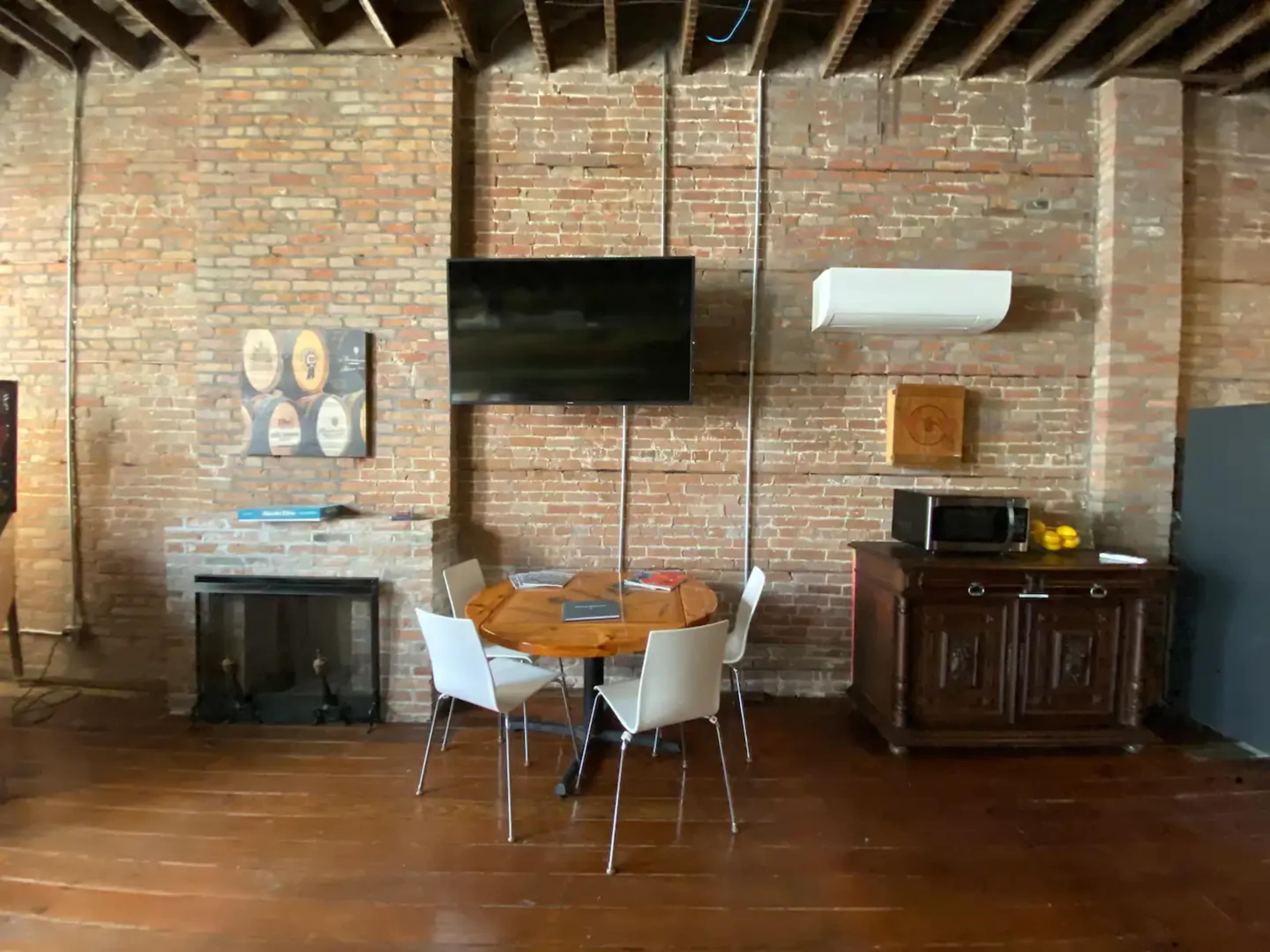 The image shows a room with exposed brick walls, featuring a round wooden table with four white chairs, a wall-mounted TV, and a wooden cabinet beside a fireplace.