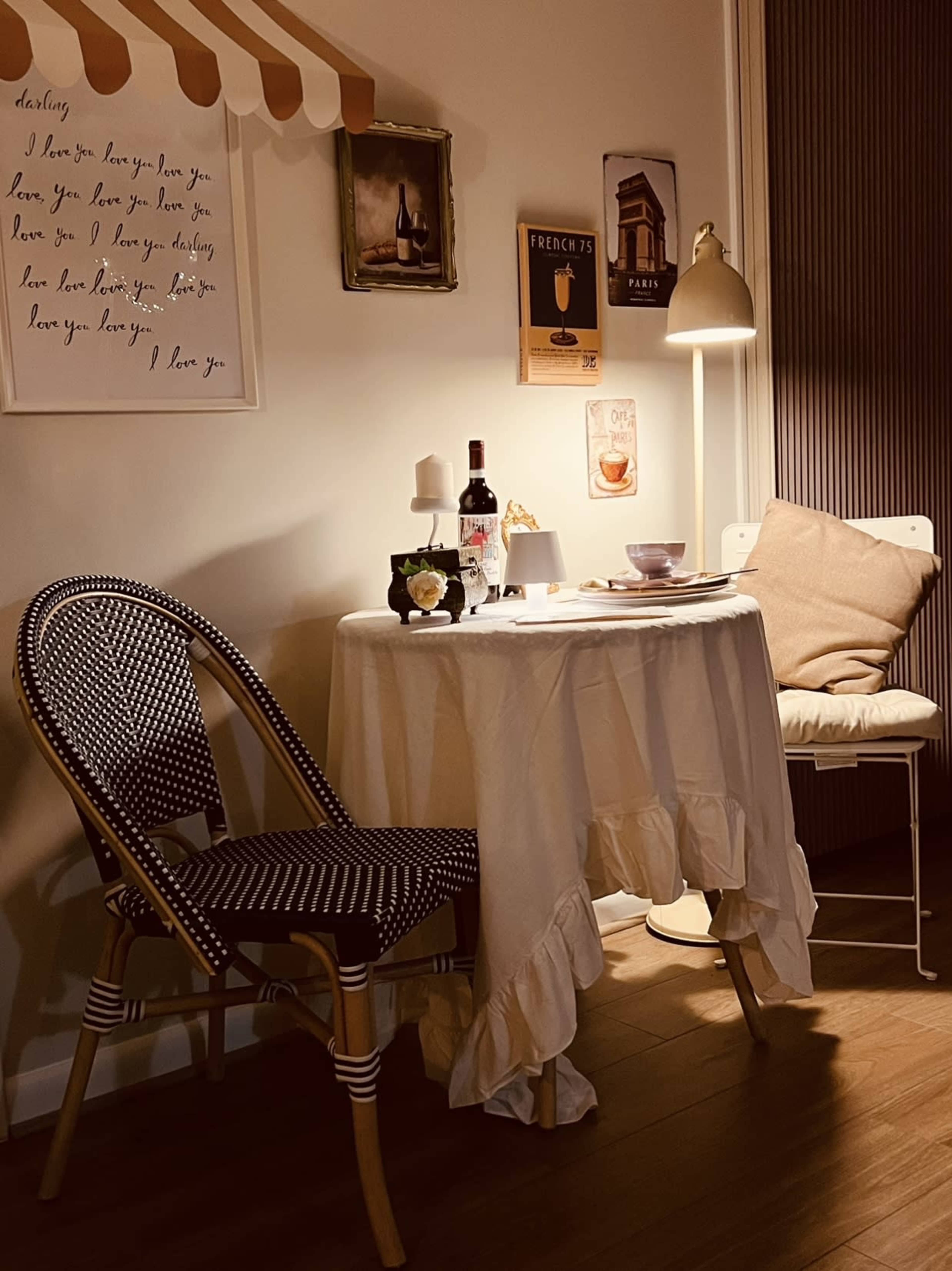A small, cozy dining space features a round table with a white tablecloth, a chair with a decorative cushion, and various decorative items, including a bottle of wine and framed artwork on the walls.